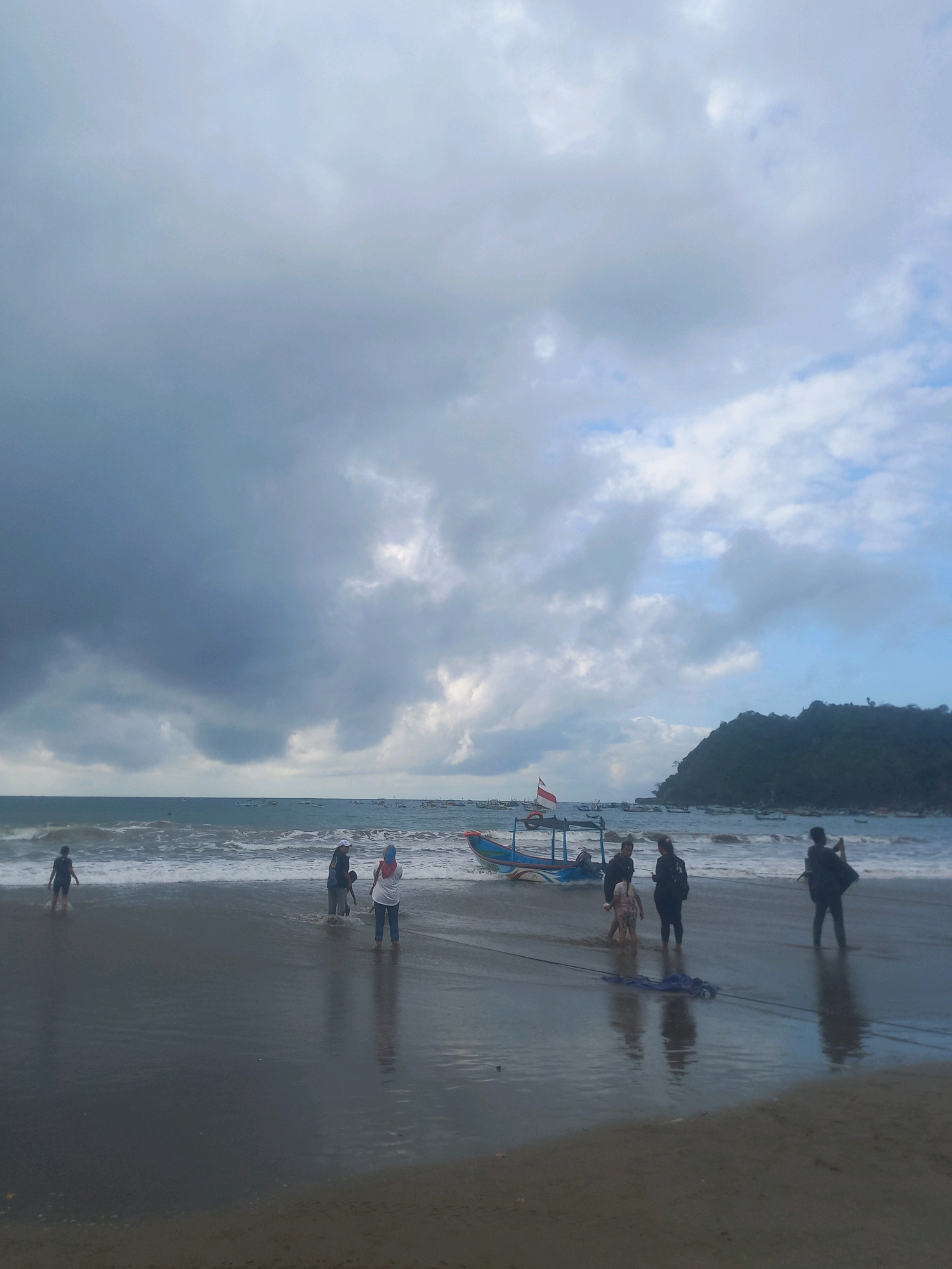 In Unknown, Indonesia, on a cloudy day at the beach, a group of people are gathered on the wet sand near the shoreline. Waves are rolling onto the beach, and several small boats are visible in the water, with one colorful boat with a red and white flag beached on the sand. A dark, hilly coastline forms the background. Several individuals are standing in the shallow water or on the sand, some facing the water, others looking towards the land or at each other. One person appears to be taking a photo with their phone. The overall scene suggests a casual outing at the beach, possibly before or after a boat activity.