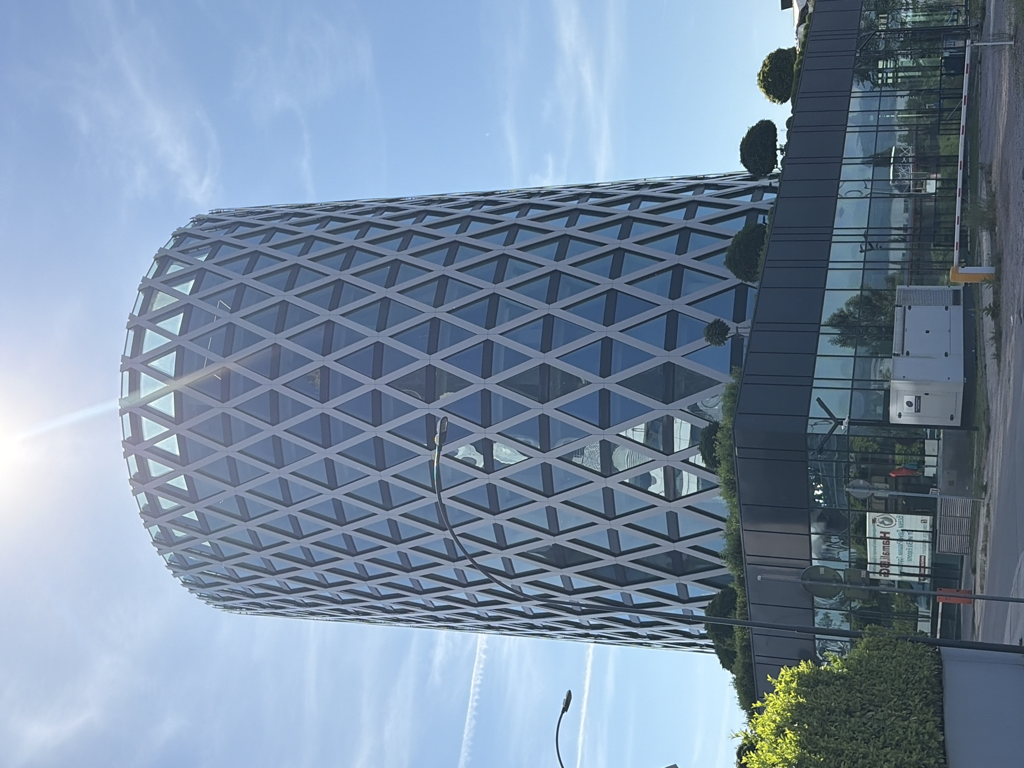 The image shows a modern, cylindrical building with a distinctive diamond-shaped facade in Sofia, Bulgaria. The building has a glass exterior with a geometric pattern of interconnected triangles formed by grey framing. The top of the building is rounded, and the sun is shining brightly, creating lens flare at the top of the image. Below the main tower is a lower, rectangular building with a glass facade and a flat roof adorned with greenery and several perfectly spherical topiary trees. In front of the lower building, a white generator is visible, along with a red and white barrier arm, suggesting an entrance or parking area. A street lamp with a curved arm is positioned to the left of the main building. The sky is clear blue with wispy white clouds and contrails from an aircraft. The scene appears to be an urban environment, likely a business district given the modern architecture.