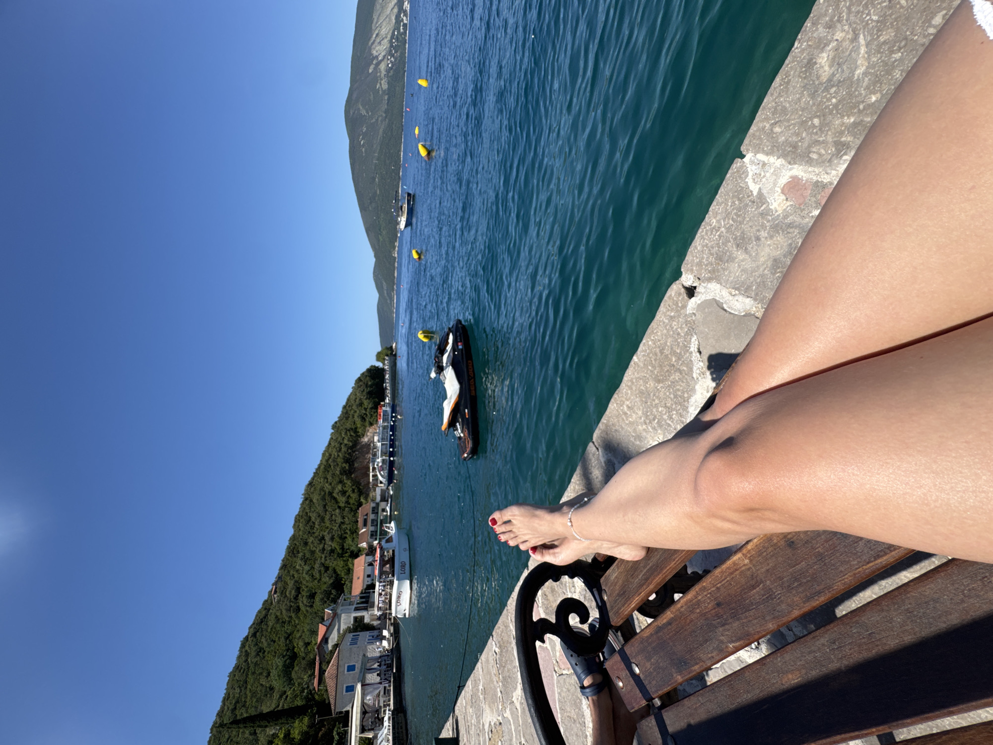 The image displays a first-person perspective of a person relaxing on a wooden slatted bench beside a coastal body of water on a clear, sunny day. The foreground prominently features the person's tanned bare legs, extended and resting on the bench. The left foot is visible with red nail polish on the toenails and a delicate white beaded ankle bracelet. Part of a dark metal armrest of the bench is visible on the left. The bench rests on a stone pier that runs along the edge of the water.
The midground shows calm, clear blue-green water. A black and white jet ski is moored near the pier on the left, and several yellow spherical buoys are floating in the water further out. Along the left shore, a small settlement of buildings with red-tiled roofs and light-colored walls, including one with distinctive blue window frames, are situated at the base of a steep, densely tree-covered green hill. Several boats are present in the bay, including a white motorboat with "LORO" visible on its side and another displaying "GRGUR" and "CROATIA".
The background is dominated by a clear, bright blue sky above distant, larger green hills and mountains that delineate the far coastline, suggesting a broad bay or a fjord-like inlet. The scene is illuminated by strong sunlight, indicating fair weather. The specific location is a coastal town in Croatia.
