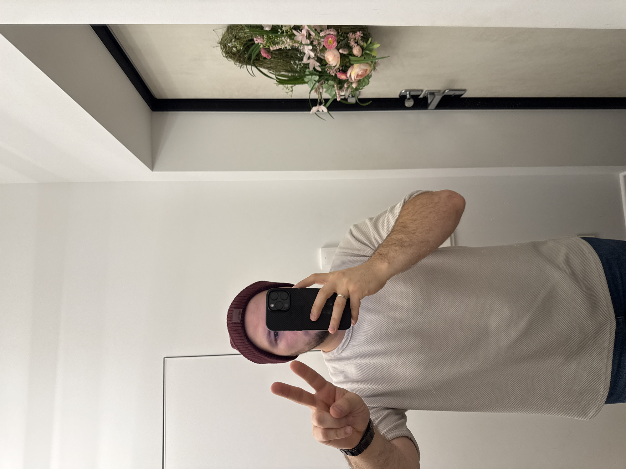 A person wearing a maroon beanie, a light-colored textured long-sleeved shirt, and jeans is holding a black smartphone in front of their face while making a peace sign with their left hand. The person appears to be taking a selfie in a mirror. The reflection shows a bouquet of pink and white flowers hanging on the ceiling above a bathroom sink with silver faucets. The room has white walls and a black trim around a recessed portion of the ceiling.
There is no evidence to support the claim that the location is Krasnoyarsk, Russia. The description will omit that claim.