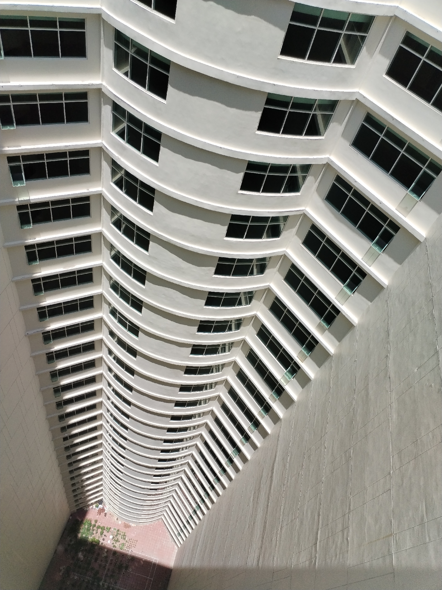 The image is a high-angle shot looking down into the courtyard of a tall, curved building. The building appears to be located in the city of Nha Trang Ward, Vietnam. The architecture features numerous windows on each floor, arranged in a consistent pattern. The exterior walls of the building are a light color. The courtyard below has a red brick surface with some vegetation growing in the corners. The overall impression is that of a modern building with a unique curved design. There are no people visible in the image. The environment appears to be sunny and bright.