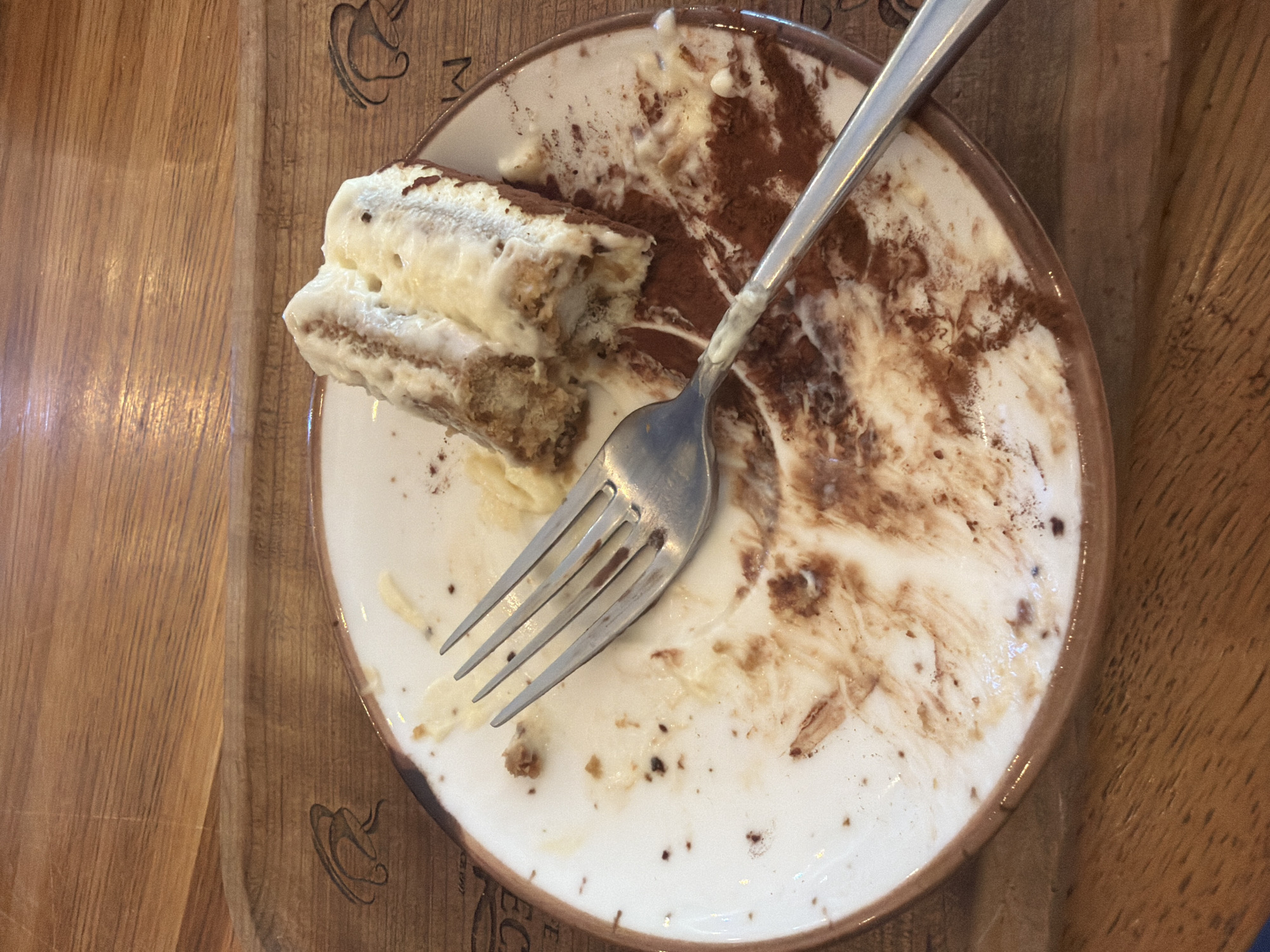 This is a top-down, close-up shot of a partially eaten piece of tiramisu on a white plate. The tiramisu has layers of sponge cake and cream, with cocoa powder dusted on top. A silver fork is resting on the plate, with some of the dessert smeared onto its tines and the plate itself. The plate is on a wooden surface, which appears to be a table or tray. There are subtle wood grain patterns and some faint etched designs on the wooden surface, including what looks like a coffee cup icon and partial letters. The scene suggests the end of a meal or dessert course. The lighting is natural and diffused, indicating daytime. There are no people visible, and the focus is on the food and the eating utensil. The context provided places this scene in Sochi, Russia.