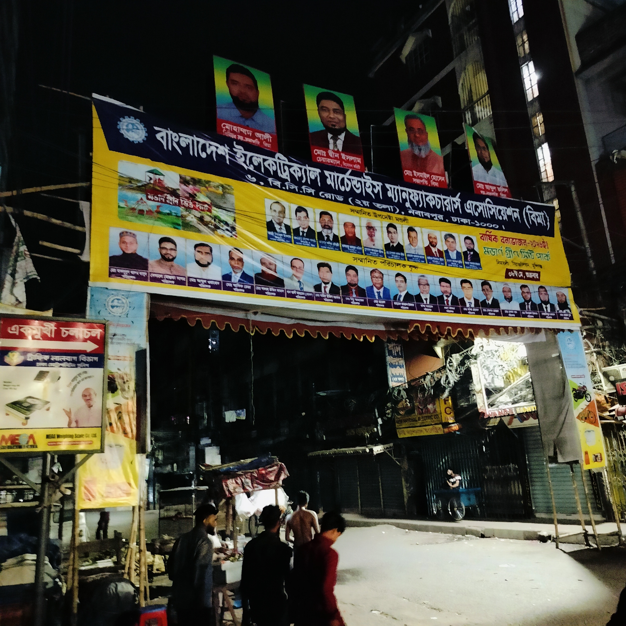 The image captures a street scene in Dhaka, Bangladesh at night. An archway spans the street, adorned with a yellow banner. This banner features the name of an organization in Bengali, "Bangladesh Electrical Merchandise Manufacturers Association (BIMA)", along with photographs of individuals identified as honored advisors and directors. Above the banner are additional portraits of men displayed vertically. The scene is illuminated by overhead streetlights and lights from nearby buildings, casting shadows. People are walking along the street, some appearing shirtless. Signs and posters are visible on the sides of the street, partially obscuring the shops. A person is standing near a cart on the street. Buildings rise in the background, and electrical wires crisscross overhead.
