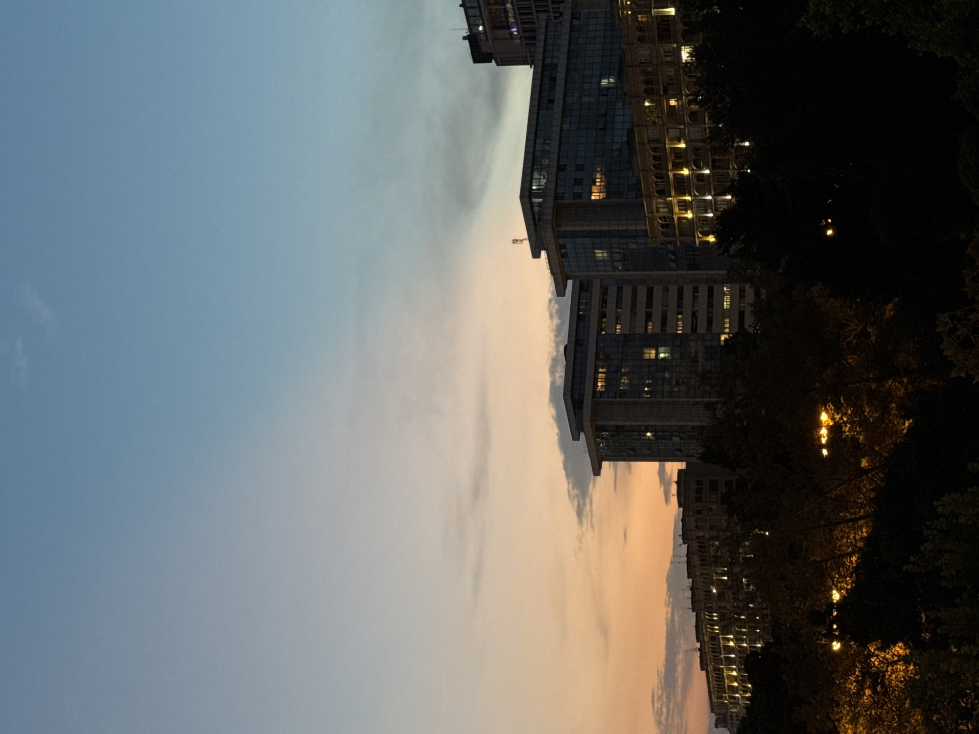 The image depicts a cityscape at dusk, with buildings and trees silhouetted against a gradient sky transitioning from orange and pink near the horizon to a clear blue overhead. Several multi-story buildings, some with modern glass facades and others with more traditional arched windows illuminated by interior lights, are visible. In the foreground, dense foliage, possibly trees, obscures the lower portion of the scene, with streetlights casting a warm glow through the branches. The architecture suggests an urban setting in Baku, Azerbaijan.