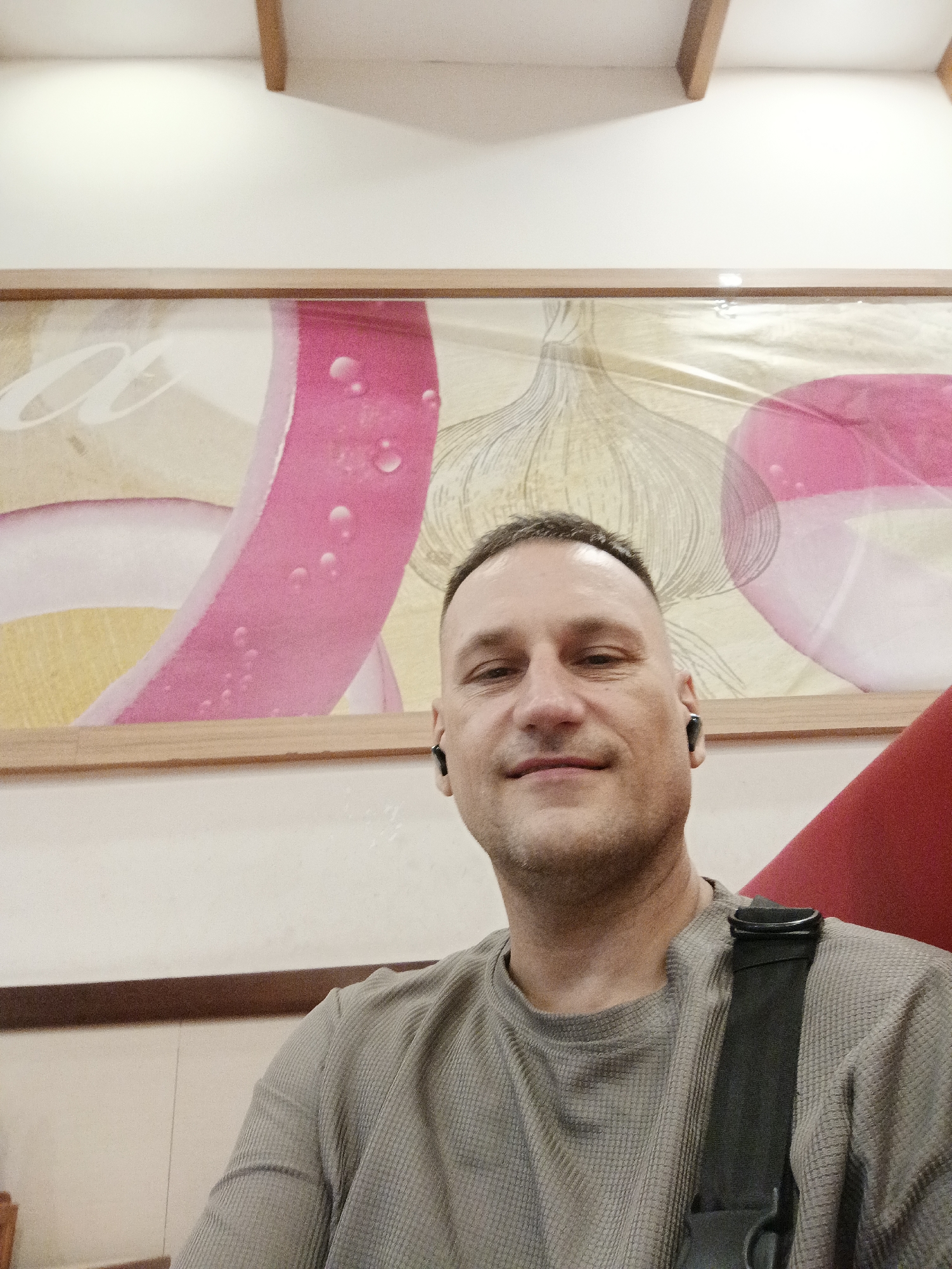 This is a medium shot, selfie-style photograph of a man with a buzz cut, wearing a grey waffle-knit long-sleeved shirt and black earbuds. He is looking directly at the camera with a slight smile. He is seated in front of a framed artwork depicting slices of red onion and garlic. The background consists of a white wall and a wooden beam across the top. A red chair is partially visible to the right of the man, and a black strap is visible across his shoulder and chest, suggesting he may be wearing a bag. The lighting appears to be indoor, possibly artificial. The overall impression is casual and relaxed. The context is Nha Trang Ward, Vietnam.