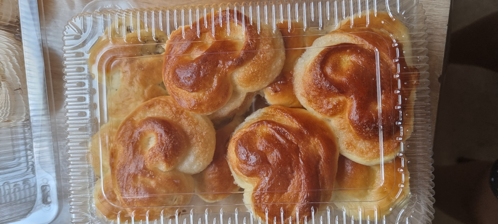 A clear plastic container holds a variety of baked goods, likely sweet rolls or pastries. The rolls are golden brown and have a swirled, knot-like shape, with some appearing to be formed into a heart shape. They are arranged in multiple layers within the container. The background appears to be a wooden surface. The image was taken in Aktobe, Kazakhstan.