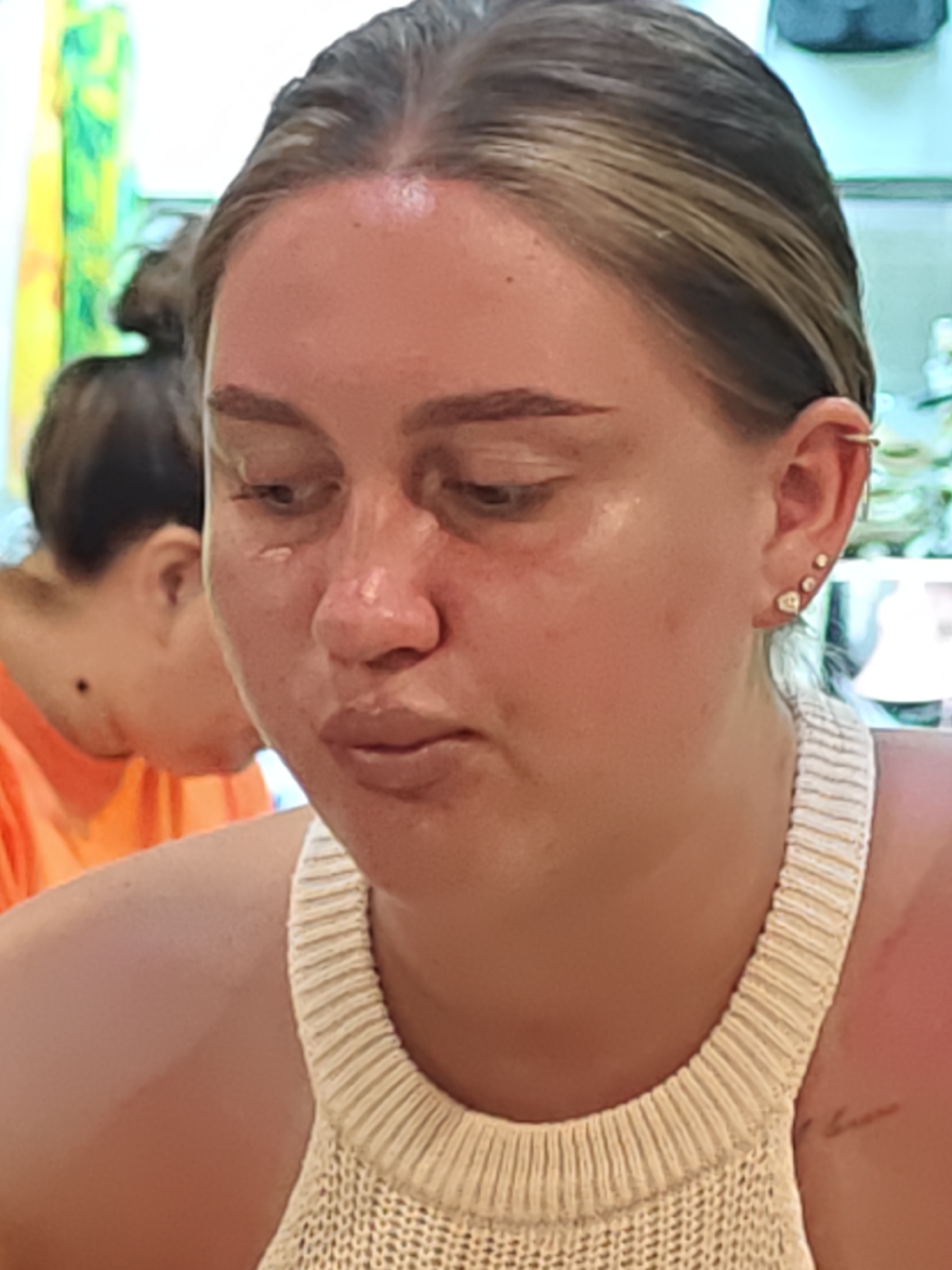 In this close-up image, a young Caucasian woman with blonde hair pulled back is the central subject, her face angled downwards and to the left. She appears to be wearing makeup, with defined eyebrows and some shine on her skin. Several small stud earrings are visible on her right ear. Her attire is a cream-colored, ribbed knit top. Behind her, another person, seen from the back, is wearing an orange shirt. The background is blurred, suggesting an indoor setting, possibly a restaurant or cafe. The image does not contain any specific landmarks or contextual cues that would allow for precise localization within Nha Trang Ward, Vietnam.