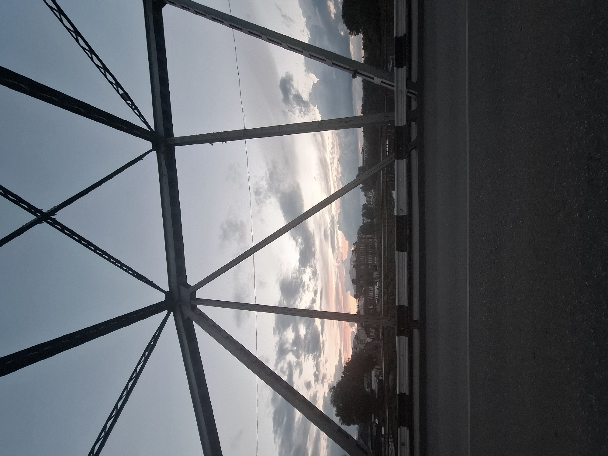 The image shows a view of a city on the horizon, seen through the latticework of a bridge, in Kungur, Russia. The sky is overcast with a sunset casting soft pink and orange hues. A multi-story building with a dark facade and many windows is visible in the center of the horizon, likely a prominent structure in the city. Trees line the banks of a river that flows through the cityscape. The bridge's metal framework is dark and industrial, framing the scene. The foreground consists of the asphalt road surface of the bridge, with a white line dividing lanes. A black and white striped barrier runs along the edge of the bridge. No people or vehicles are clearly visible. The overall impression is a tranquil, somewhat moody, urban landscape at dusk.