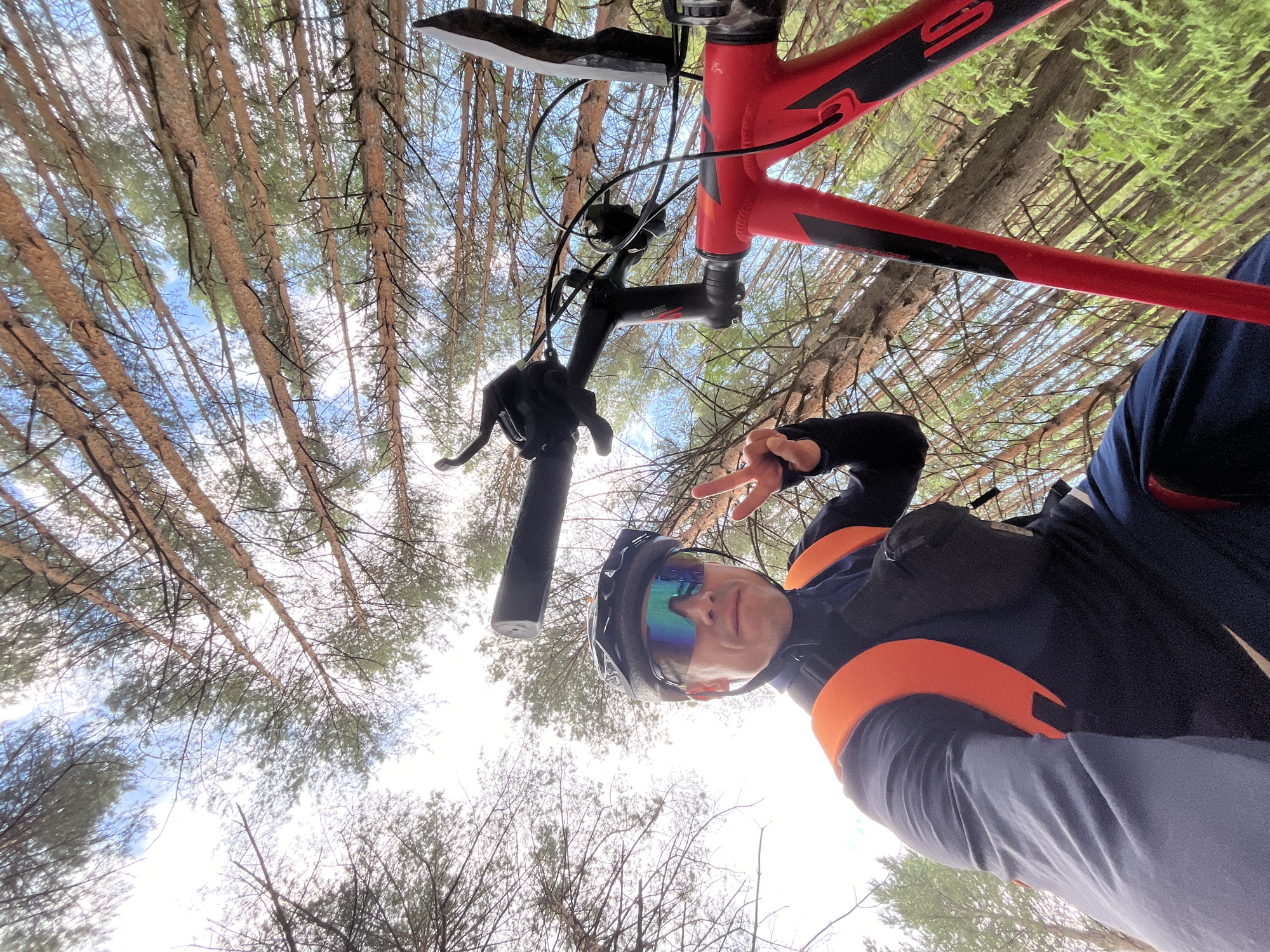 The image is a low-angle, wide shot of a person on a red bicycle in a forest. The person, wearing a black helmet, blue long-sleeved shirt, orange backpack, and sunglasses, is giving a peace sign with their left hand. The bicycle's handlebars and front frame are visible. The background consists of tall trees with a blue sky peeking through the branches. The scene appears to be a recreational setting. The location is not specified in the image.