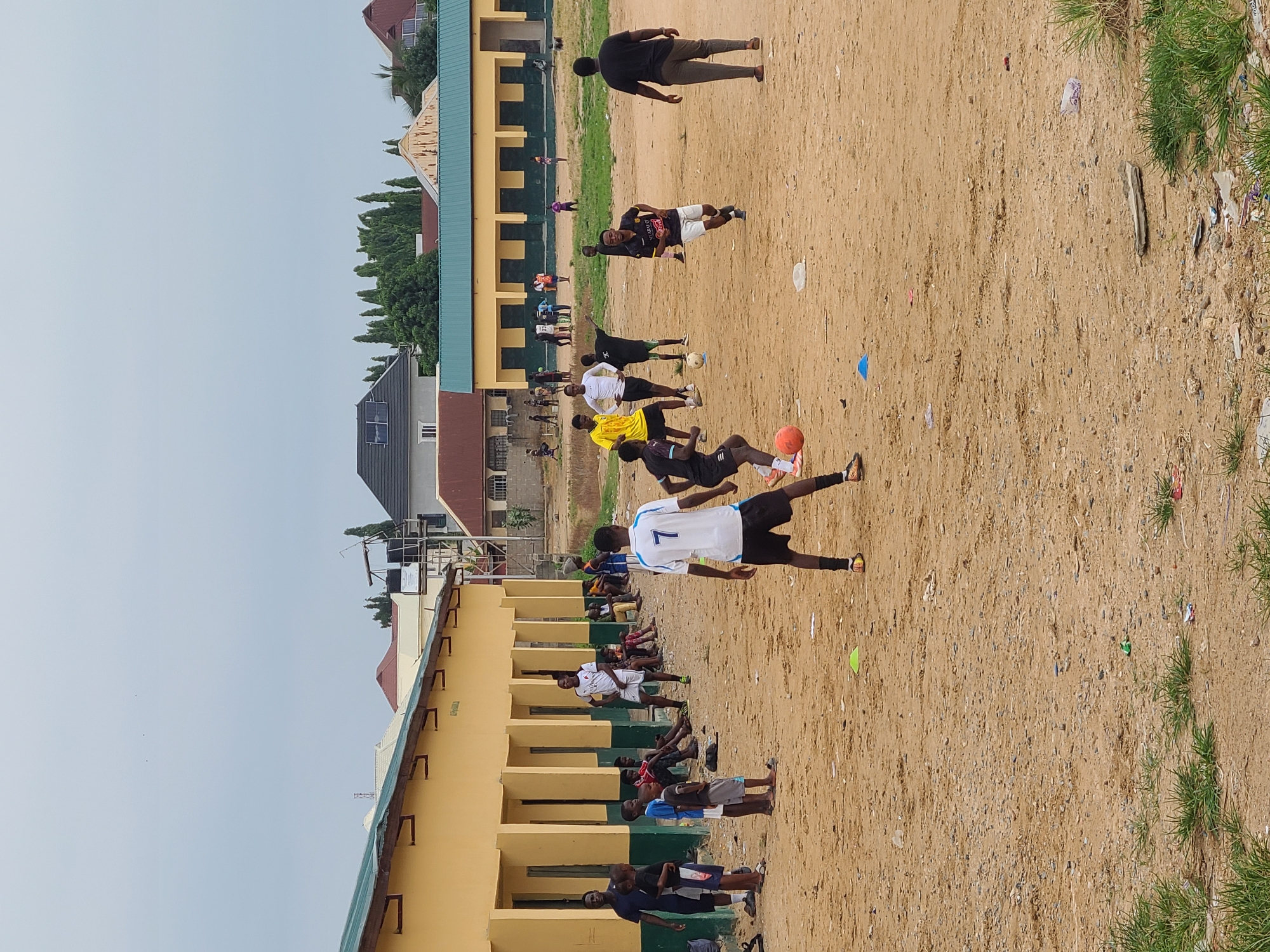 In Masaka, Nigeria, a group of young men and boys are engaged in a football match on a dirt field. The field is uneven and littered with debris. In the background, a long, single-story building with a yellow facade and a green roof stands prominently. The building has multiple doorways and appears to be a school or community center. Several spectators are seated on the ground under the building's overhang. The sky is overcast, suggesting a cloudy day. The overall scene depicts a casual sporting activity in a somewhat rudimentary urban setting.