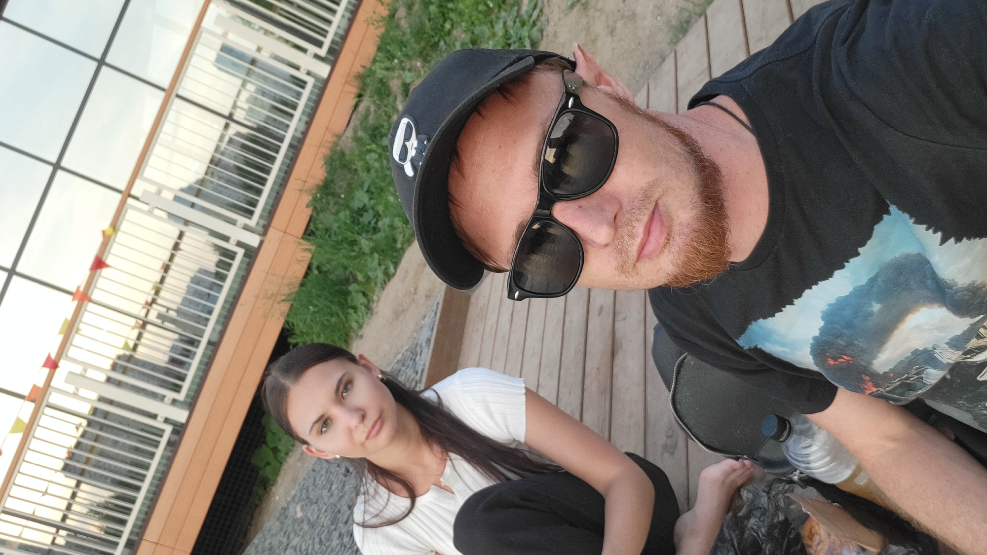 Two individuals are seated on a wooden deck outdoors. A woman with long dark hair is seated to the left, wearing a white top and black pants. A man with a red beard, sunglasses, and a black baseball cap is seated to the right, wearing a black t-shirt with a graphic print. A backpack and a water bottle are visible on the deck between them, along with a plastic bag containing food items. In the background, a building with large windows and railings is visible, adorned with small triangular flags. The overall setting suggests a casual outdoor gathering.