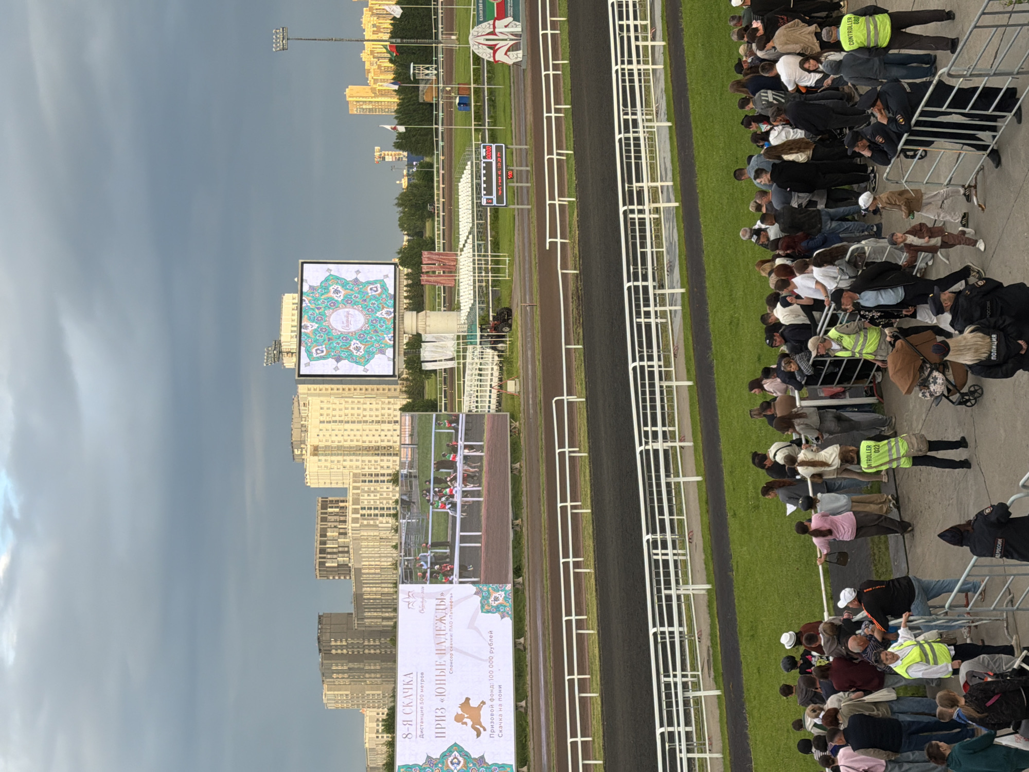This is a wide shot of a horse race track from a high angle. In the foreground, a crowd of people are gathered behind a metal fence. Several individuals are wearing high-visibility vests, indicating they are staff or security. The crowd appears to be watching an event on the track. In the middle ground, the race track itself is visible, with a black dirt surface and white railings. Beyond the track, there are rows of empty white spectator seats and some structures associated with a racecourse. A large digital billboard displays an image of a horse and text in Russian. In the background, a cityscape with modern high-rise buildings is visible under a cloudy sky. The overall atmosphere suggests an outdoor event at a sports venue.