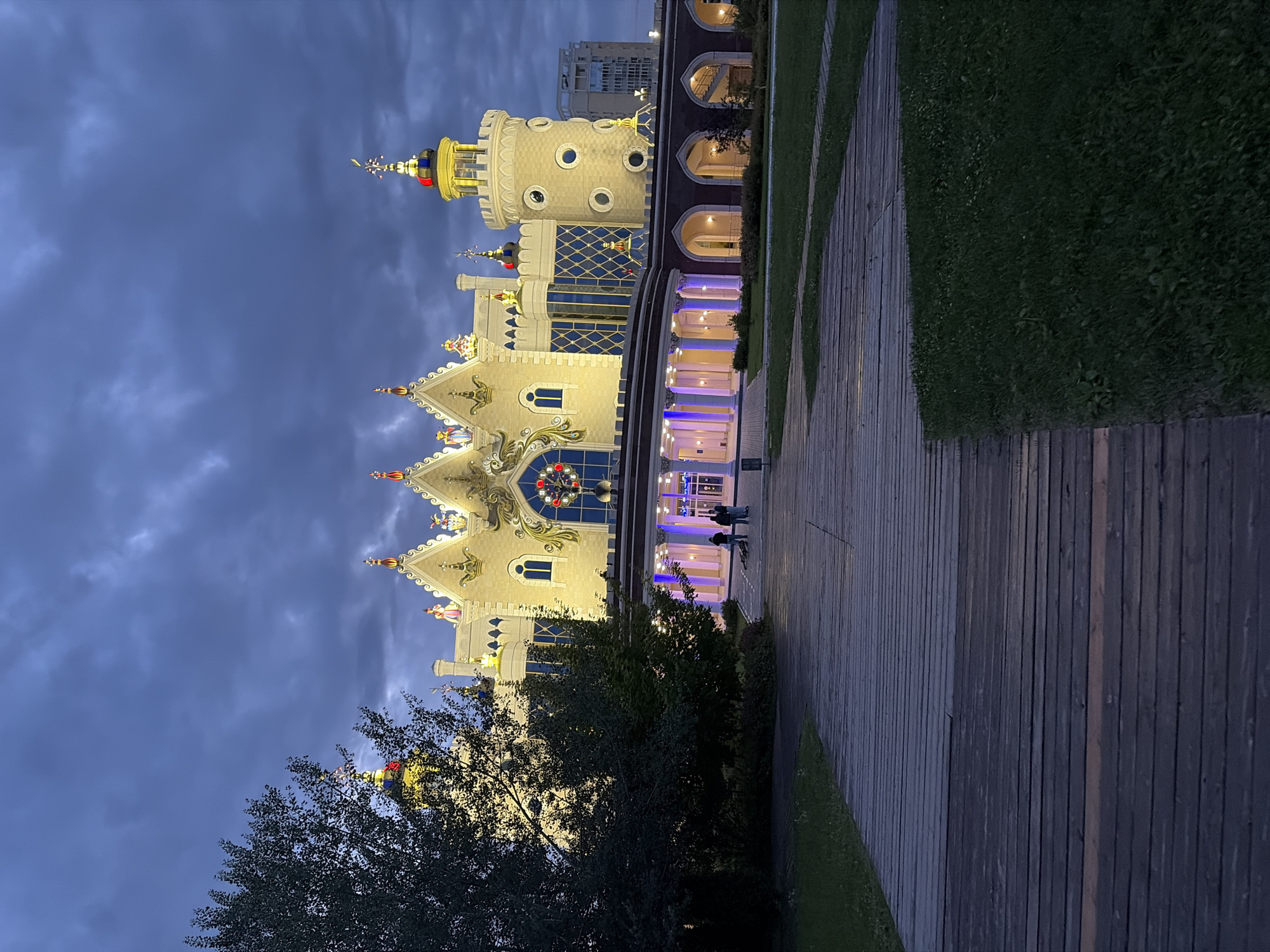 A whimsical, fairytale-like building with multiple towers and ornate decorations is illuminated against a dusky sky. The building, likely a tourist attraction given its elaborate design, features a mix of pale yellow and dark red accents, with a prominent round tower topped by a gold and red spire. The facade is adorned with decorative gables, spires, and what appear to be figures or statues. Large arched windows and doorways are visible, with interior lights casting a warm glow.
A paved walkway made of wooden planks runs alongside the building, bordered by a neatly trimmed green lawn. Several silhouetted figures can be seen walking on the pathway near the entrance of the building. The time of day is twilight, as indicated by the dark blue sky and the artificial lighting on the building and pathway. The weather appears calm with some cloudy textures in the sky. The scene evokes a sense of enchantment and wonder, typical of a fairy tale setting. There is no visible text in the image. The location context provided suggests this is in Kazan, Russia.