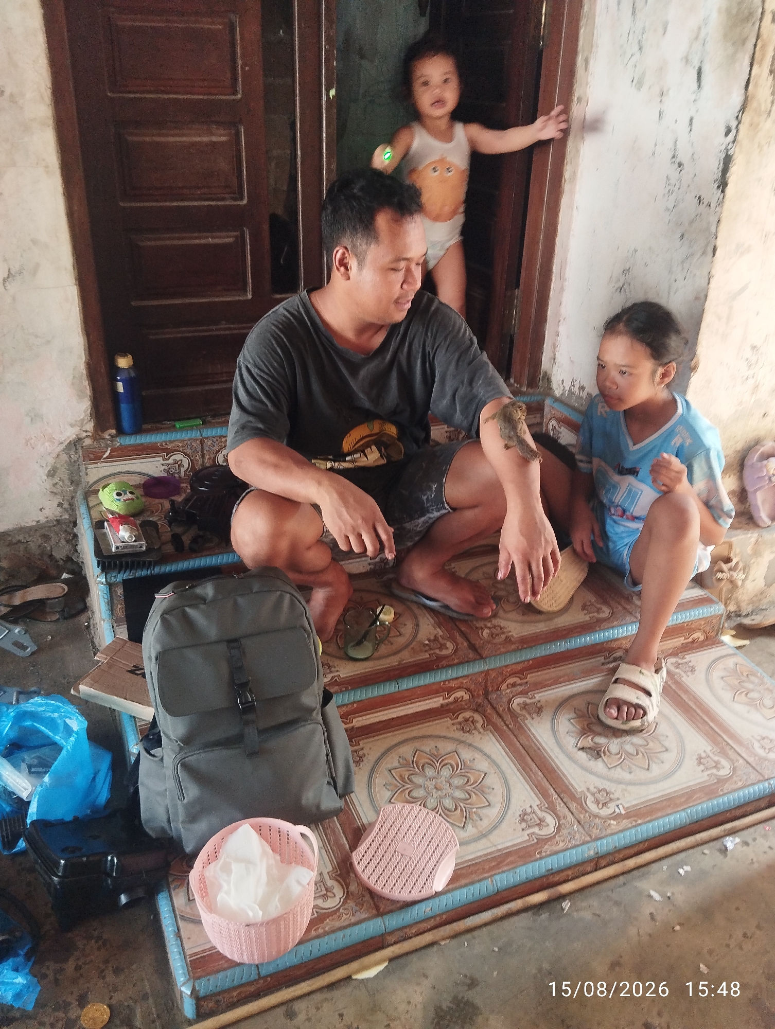 In Tegal Rejo, Indonesia, an image captures a father and his two children. The father, a man with short black hair, wears a dark gray t-shirt and dark shorts. He is seated on a tiled step, his left arm resting on his knee, and appears to be holding something small and furry, possibly a lizard or gecko, on his arm. To his right sits a young girl with dark hair, wearing a light blue t-shirt with white and blue graphics and blue shorts. She is looking towards the left. Behind them, standing in a doorway, is a young child, a boy or girl, wearing a white tank top with an orange cartoon image and white bottoms, with arms outstretched. The environment appears to be an outdoor space with a weathered wall on the right and a wooden door on the left. Various objects are present, including a gray backpack, a pink woven basket filled with white tissues, blue plastic bags, a flip-flop, and a bottle. The tiled steps are decorated with floral patterns in brown and blue. A timestamp in the lower right corner reads "15/08/2026 15:48".