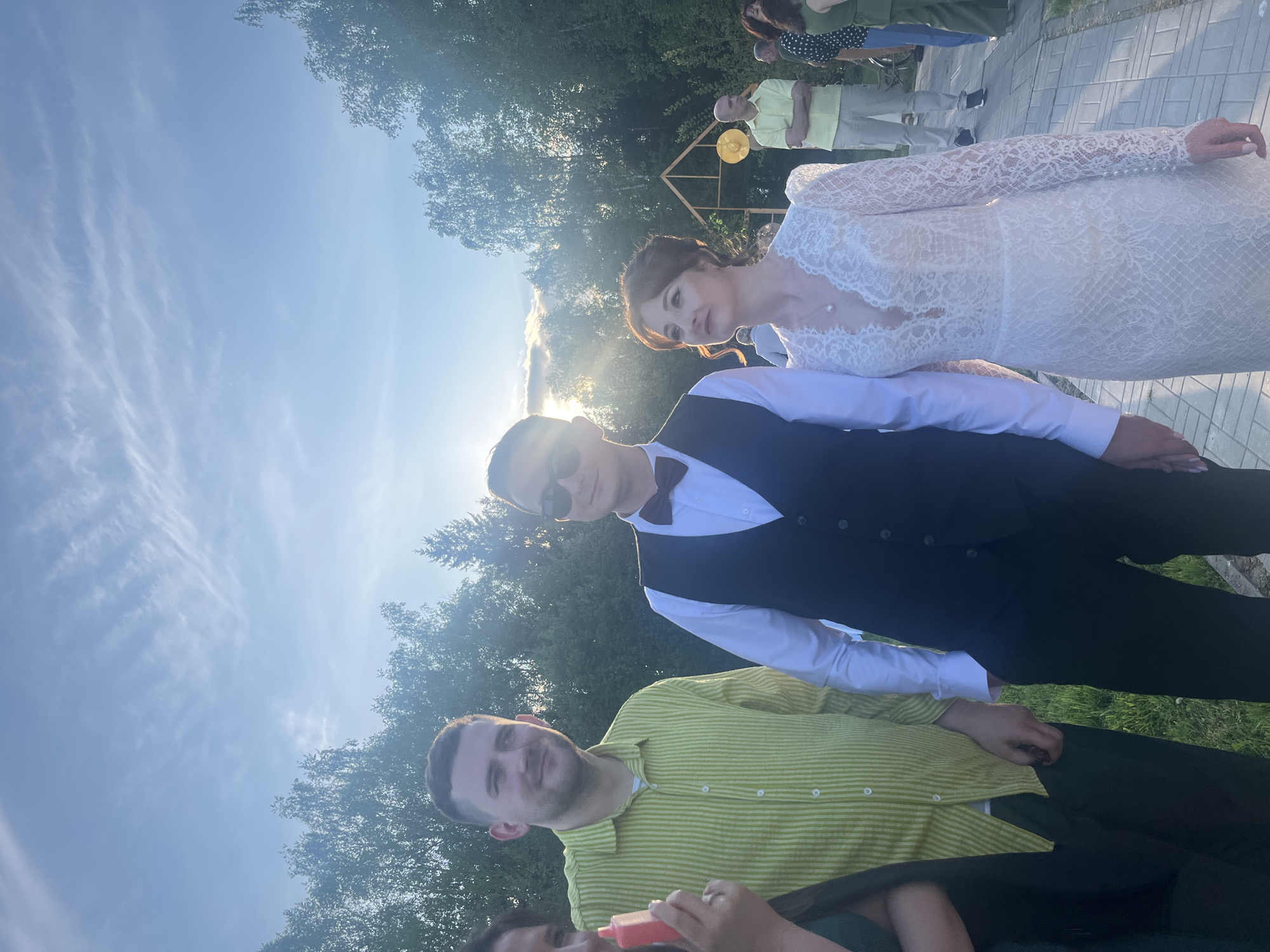 A photograph taken in the city of Алопово, Russia, depicts a wedding party. The bride, in a white lace gown, stands beside a man wearing a bow tie and vest. Another man in a striped yellow shirt is visible at the bottom of the frame. In the background, other people are seen near a structure decorated with a yellow balloon. The setting appears to be outdoors, with trees and a bright, sunny sky.
