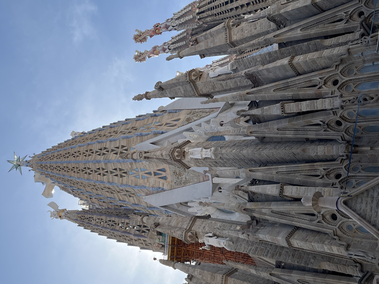 An upward-looking shot captures the intricate, towering spires of the Sagrada Familia basilica in Barcelona, Spain, under a clear blue sky with a few wispy clouds. The perspective emphasizes the immense height and detailed artistry of the structure.
The central focus is one of the main spires, adorned with a large, faceted star at its apex, glinting in the sunlight. This spire features a textured, geometric pattern in light stone and blue accents, resembling stained glass windows. To the right, multiple other spires rise, showcasing complex Gothic and Art Nouveau-inspired stonework, pointed finials, and decorative elements. Some of these spires have small, colorful, fruit-like sculptures at their very top, partially visible against the sky.
The stonework throughout the visible sections of the basilica is highly detailed, with ornate carvings, rounded windows, and varying textures. Towards the bottom-right, some orange scaffolding is visible, indicating ongoing construction or maintenance, a common sight at the Sagrada Familia. The bright sunlight suggests it is daytime, casting some shadows and highlighting the architectural contours. No discernible text is visible in the image.