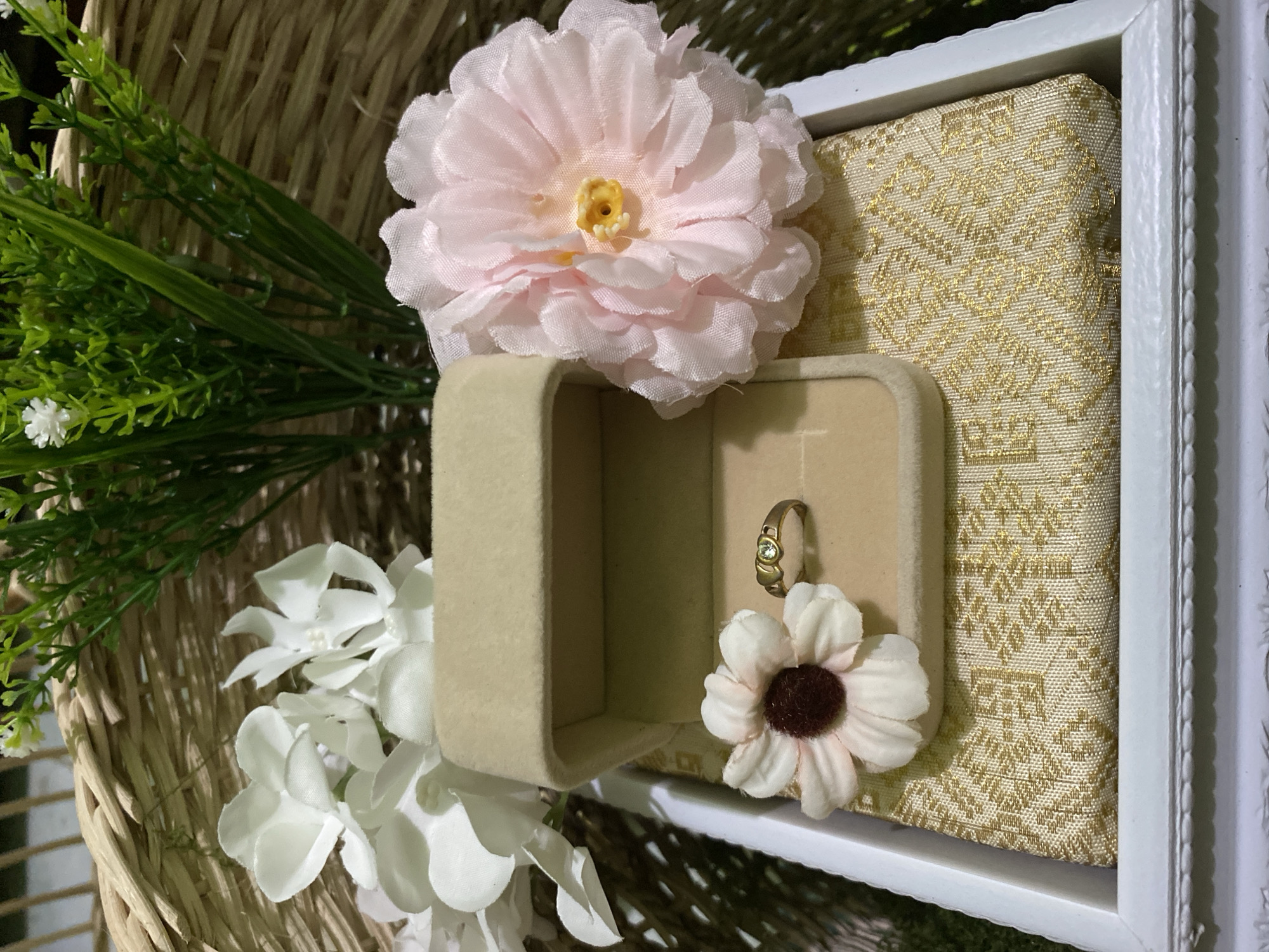 This media file showcases a close-up, overhead view of a decorative ring box and a ring, likely presented as part of a wedding or engagement ceremony. The scene is set within a woven basket, hinting at a natural or rustic aesthetic.
The primary subjects are a beige, velvet-lined ring box, open to reveal a single gold ring with a small, clear gemstone. Beside the open box, a small, cream-colored artificial flower with a dark brown center is placed. To the left of the ring box and basket, a larger, light pink artificial flower with a yellow center is prominently featured. Interspersed with these floral elements are artificial green sprigs and delicate white flowers.
The ring box itself rests on a gold and cream patterned fabric, possibly silk or brocade, which is enclosed within a white decorative frame. This framed element suggests the presentation is part of a more elaborate setup, perhaps a wedding gift or display. The overall impression is one of elegance and thoughtful arrangement, focusing on the symbolism of commitment and love.
There is no indication of time of day, weather, or specific location cues beyond the basketry and floral elements, which are common in celebratory decorations. No text is visible in the image. The composition is focused and intimate, highlighting the jewelry and decorative elements.