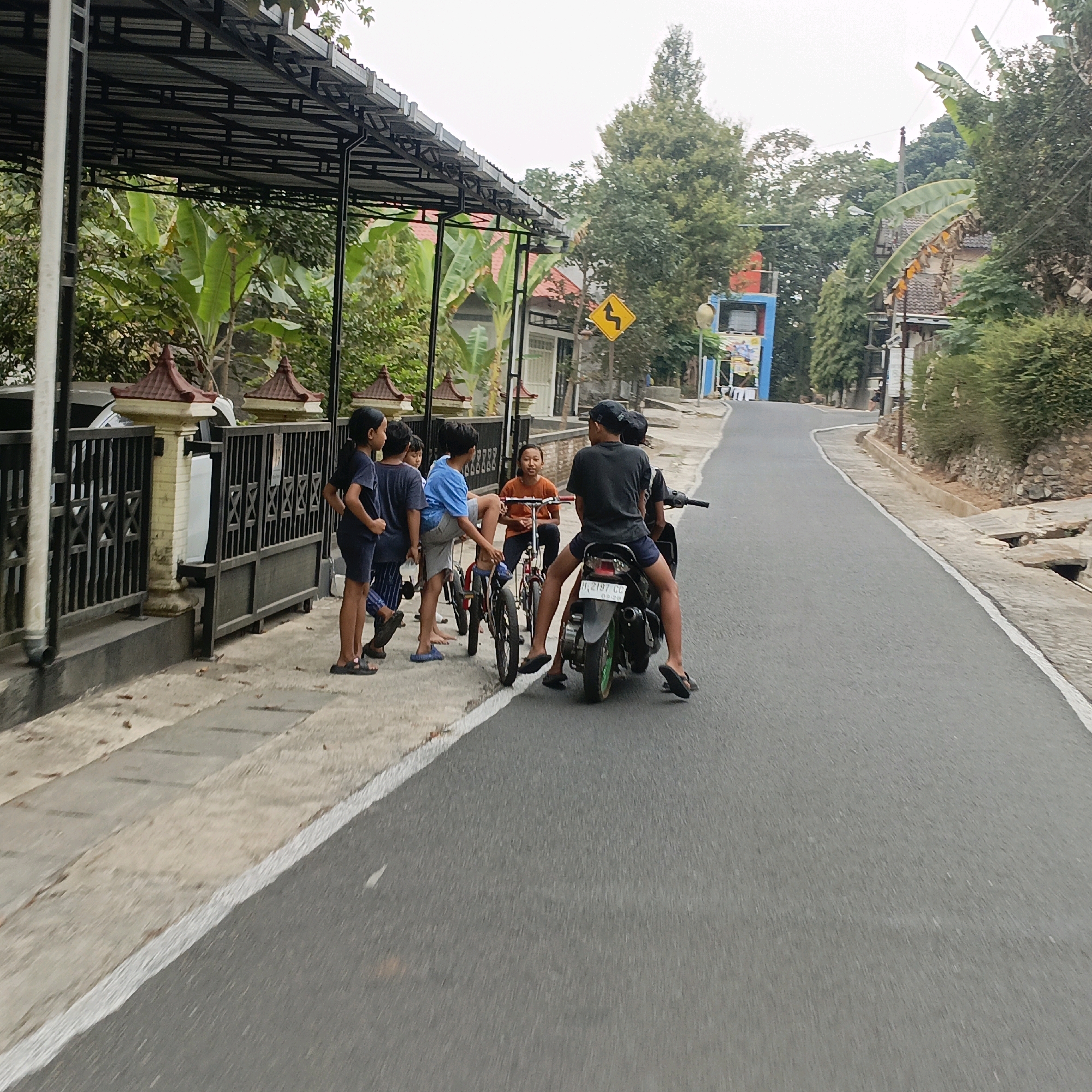 The image depicts a street scene in Salatiga, Indonesia, with a group of children on a sidewalk next to a road. On the sidewalk, there are seven children. Four children are standing near a fence, and three children are near bicycles. One child is sitting on a bicycle, and two children are standing near their bicycles. On the road, two children are on a motorcycle. One child is driving the motorcycle, and the other child is sitting behind them. The background shows buildings, trees, and a cloudy sky. The architecture suggests a residential area. The overall mood of the image is casual and everyday, capturing a moment of childhood activity in a local Indonesian setting.
