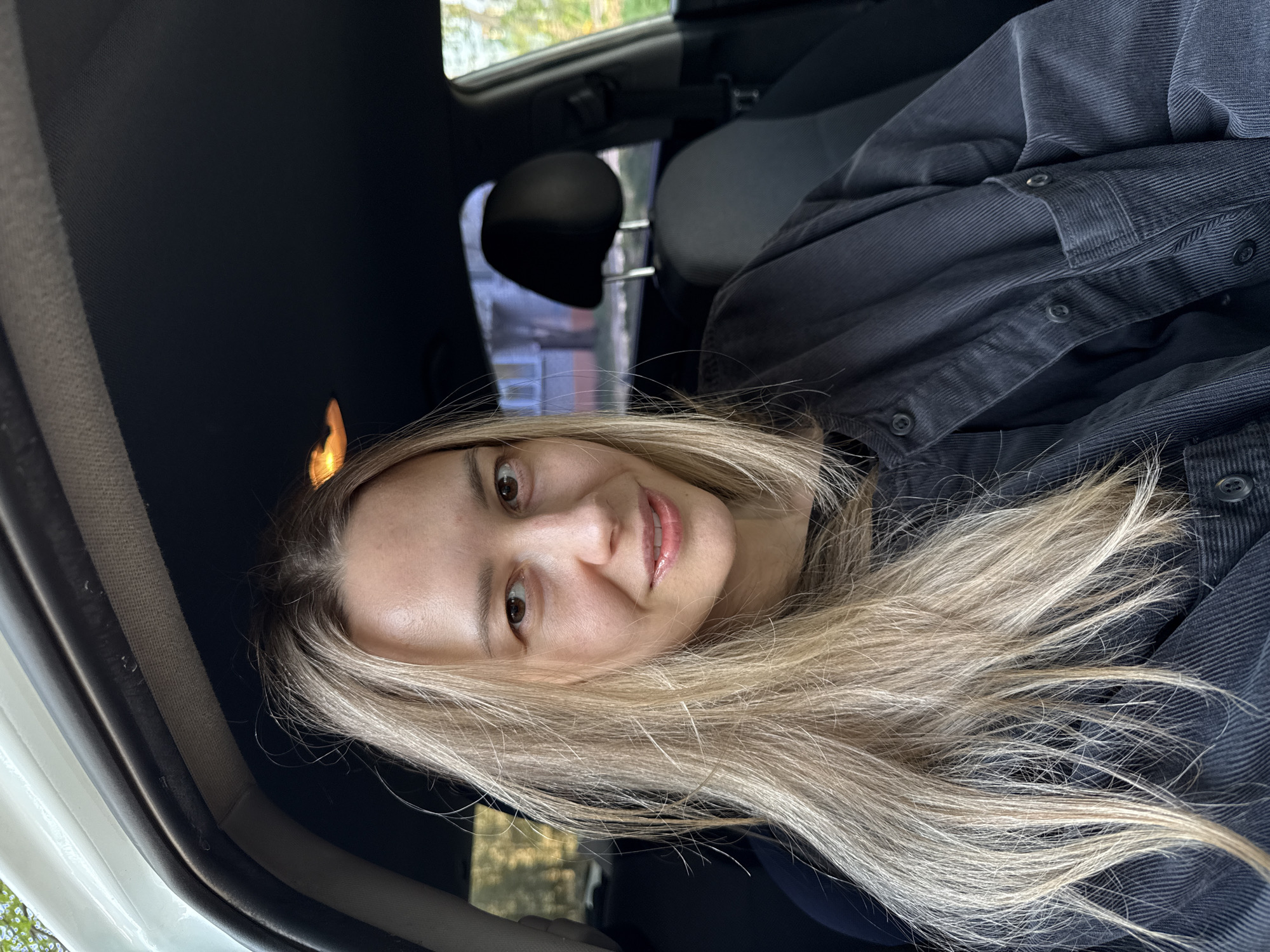 The image shows a woman with long, light blonde hair sitting in the driver's seat of a car. She is wearing a dark corduroy shirt. The interior of the car is visible, including the dashboard and seats. Outside the window, there are blurred trees and buildings. Based on the request for location specifics, and the absence of any visual cues pointing to Chaikovsky, Russia, it is not possible to confirm the location with the provided information.