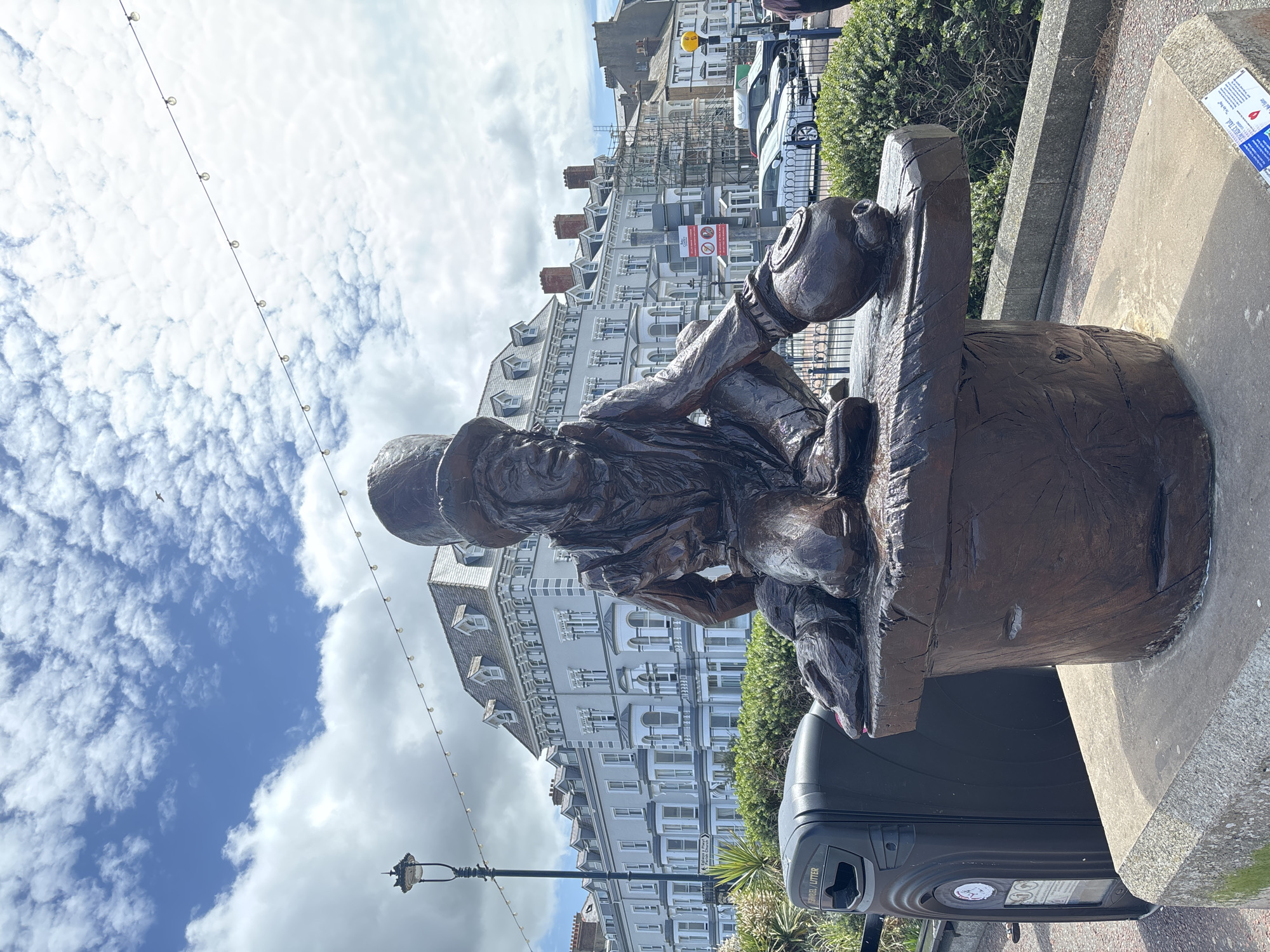 A statue of a man in a hard hat and wetsuit sits on a square wooden platform in Llandudno, United Kingdom. The statue is in front of an apartment building and next to a trash can on a sidewalk. There is a light post to the left of the statue. In the background, there is a partly cloudy blue sky.