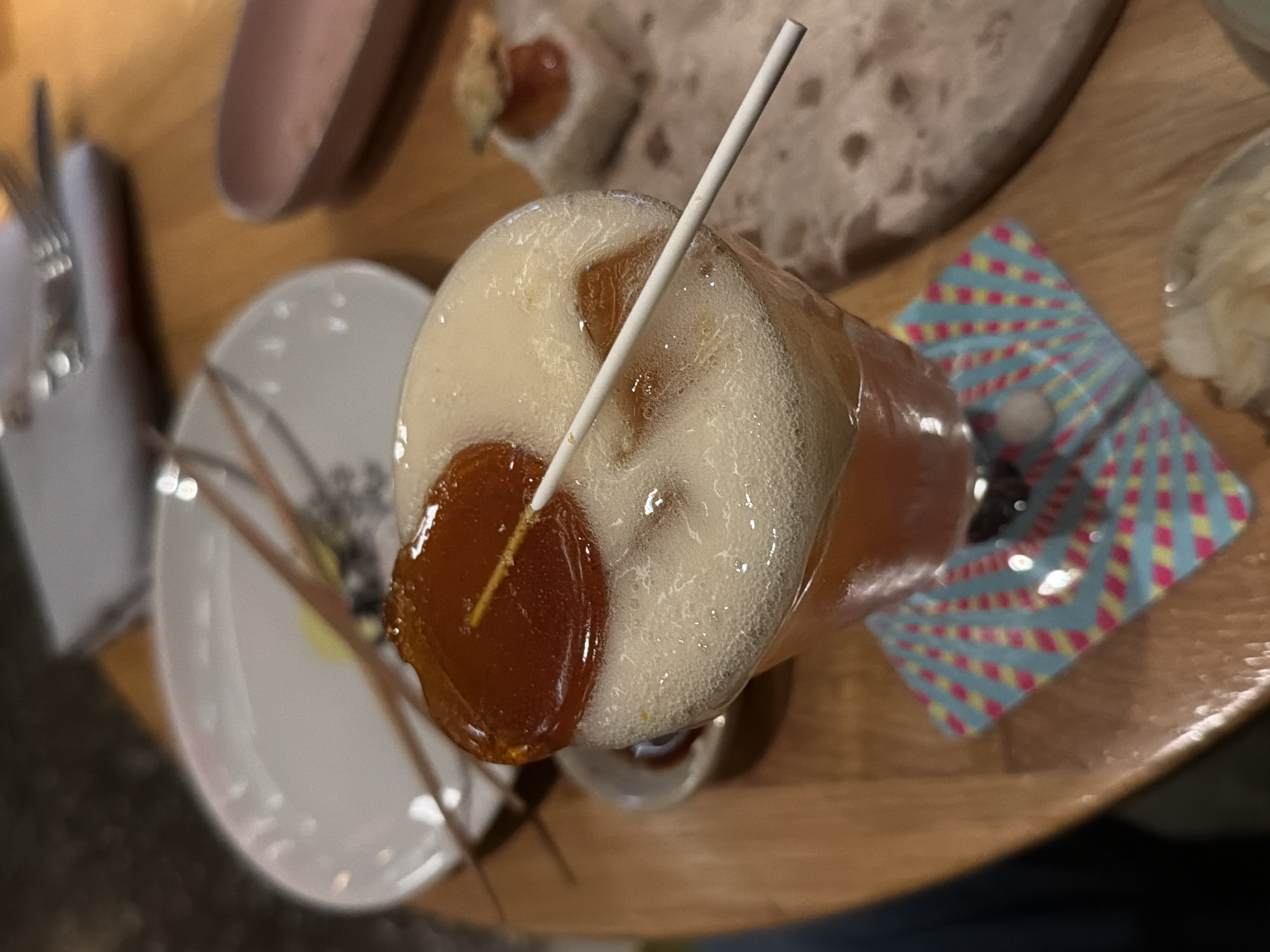 The image presents a high-angle, slightly tilted view of a beverage and accompanying items on a wooden table, likely within a restaurant or bar setting in Belgrade, Serbia.
The central subject is a tall glass filled with an amber-colored, effervescent drink, possibly a beer or cocktail, topped with a thick, creamy layer of white foam. A white straw is inserted vertically into the beverage. A prominent garnish, appearing as a glossy, deep orange, translucent candied item (resembling a lollipop or preserved fruit slice) on a thin stick, rests partially submerged on the foam, adding a vibrant touch.
The glass is placed on a colorful coaster, which features a radiant pattern of alternating blue, pink, and yellow stripes. To the left, a small white ceramic plate holds a set of metal cutlery, including forks. In the upper background, a light-colored, irregularly shaped plate or tray, possibly made of stone or textured pottery, holds a small, blurry food item, suggesting an appetizer or snack. Further blurry elements in the background hint at more dishes or table settings.
The lighting is dim, consistent with an indoor, possibly evening, ambiance. There are no visible people, discernible specific activities beyond dining, or any readable text in the image. The scene conveys a relaxed atmosphere typical of a dining establishment.