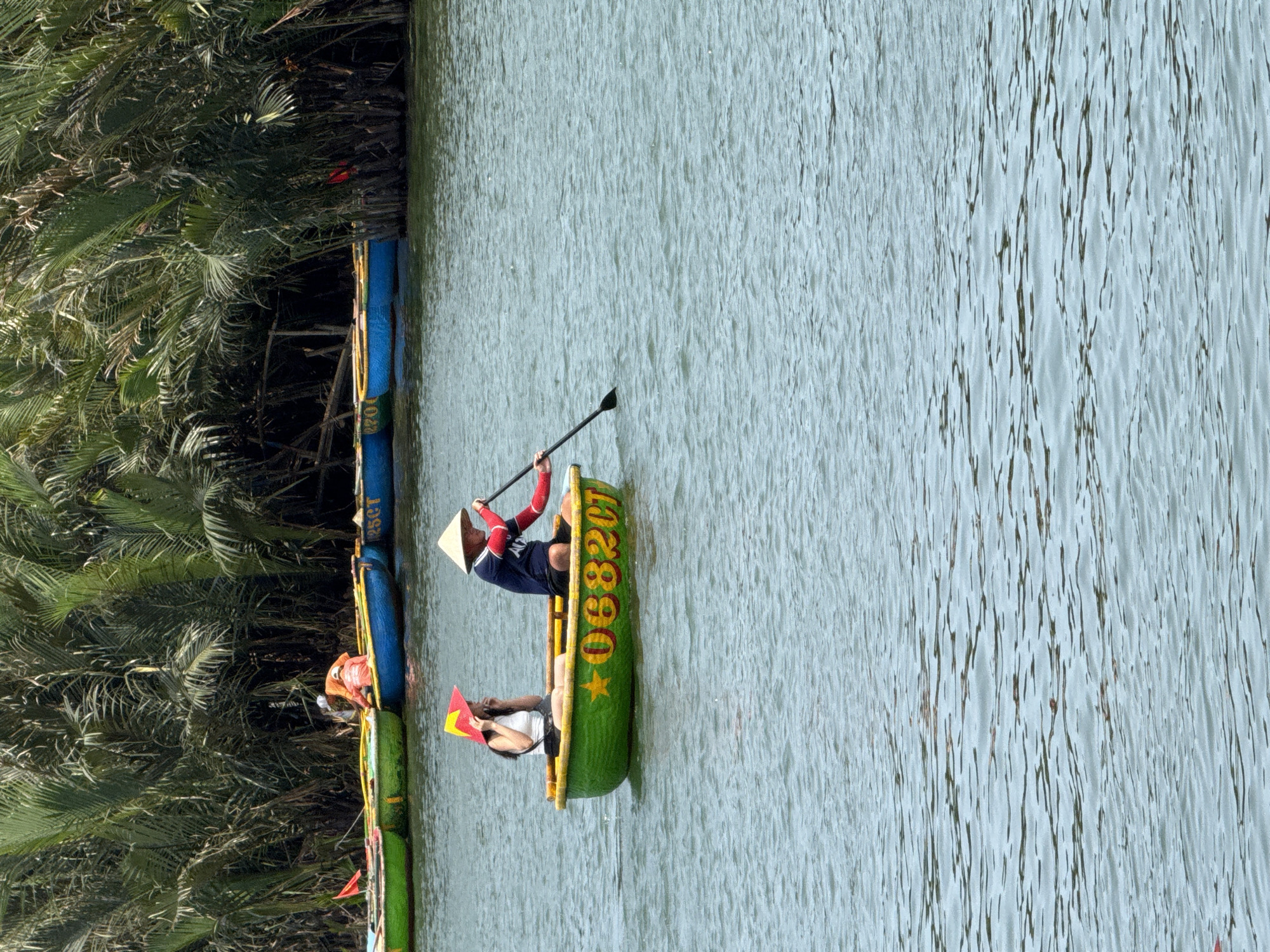 The image, rotated vertically, depicts a tranquil scene on a river or canal, likely in Đà Nẵng, Vietnam. The main subject is a person wearing a traditional Vietnamese conical hat (nón lá) and a dark blue long-sleeved shirt with red armbands, actively paddling a green basket boat (thúng chai) with a long oar. Seated in the same boat, facing away from the paddler, is another individual wearing a white top, holding a small red flag emblazoned with a yellow star – the national flag of Vietnam.
The green basket boat has yellow trim and features the numbering "06820T" and a yellow five-pointed star painted on its side. The water is calm with subtle ripples, reflecting a light blue-green hue. The left bank of the waterway is densely covered with lush green tropical foliage, appearing to be palm trees or similar vegetation. Further along the bank, other colorful basket boats are visible; one blue boat prominently displays the text "125GT" vertically on its side. A third person, wearing an orange life vest, is partially visible in another boat near the bank. The bright, natural lighting indicates it is daytime, suggesting a pleasant and peaceful environment suitable for tourism and recreational activities like a traditional boat tour.