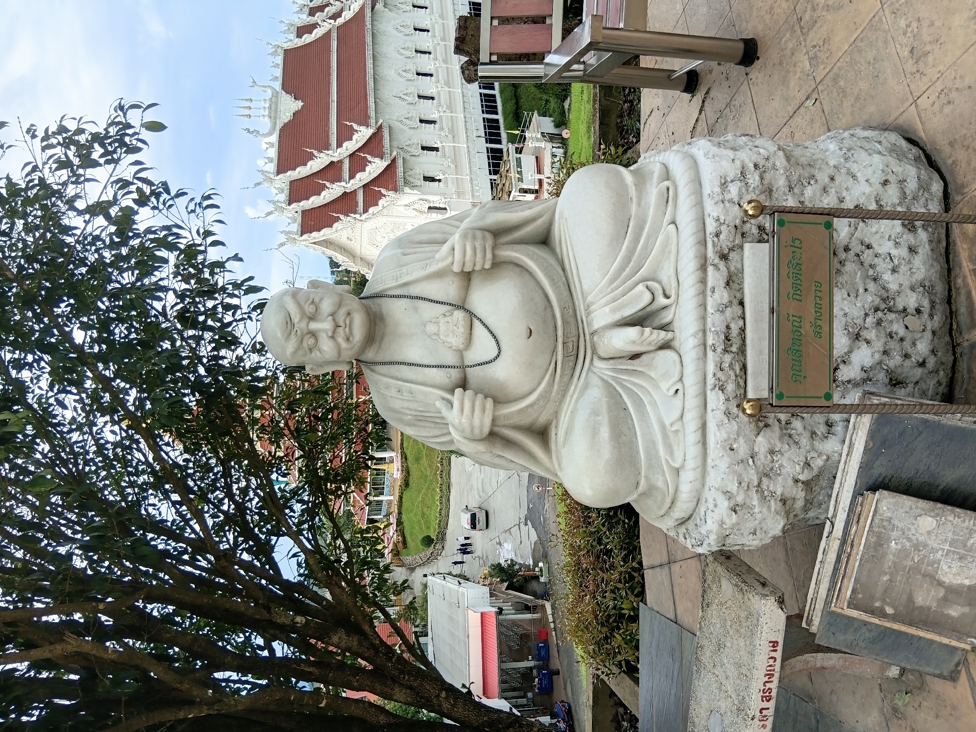 The image shows a marble statue of a seated, overweight figure in a lotus position, wearing a necklace with a small Buddha pendant. The statue is on a rough, natural-looking stone base. Behind the statue, a large white temple with red tiled roofs and intricate white architectural details is visible. To the left of the statue, a large tree with dark bark and green leaves partially obscures the view of the temple. In the middle ground, there are several buildings with red accents, a paved area with a white van and a few people walking, and a grassy slope. The foreground shows a paved area with some stacked stone slabs and a sign with Thai script. The scene appears to be outdoors in a temple complex or cultural site, possibly in บ้านเหล่าพัฒนา, Thailand, given the architectural style and the Thai script.