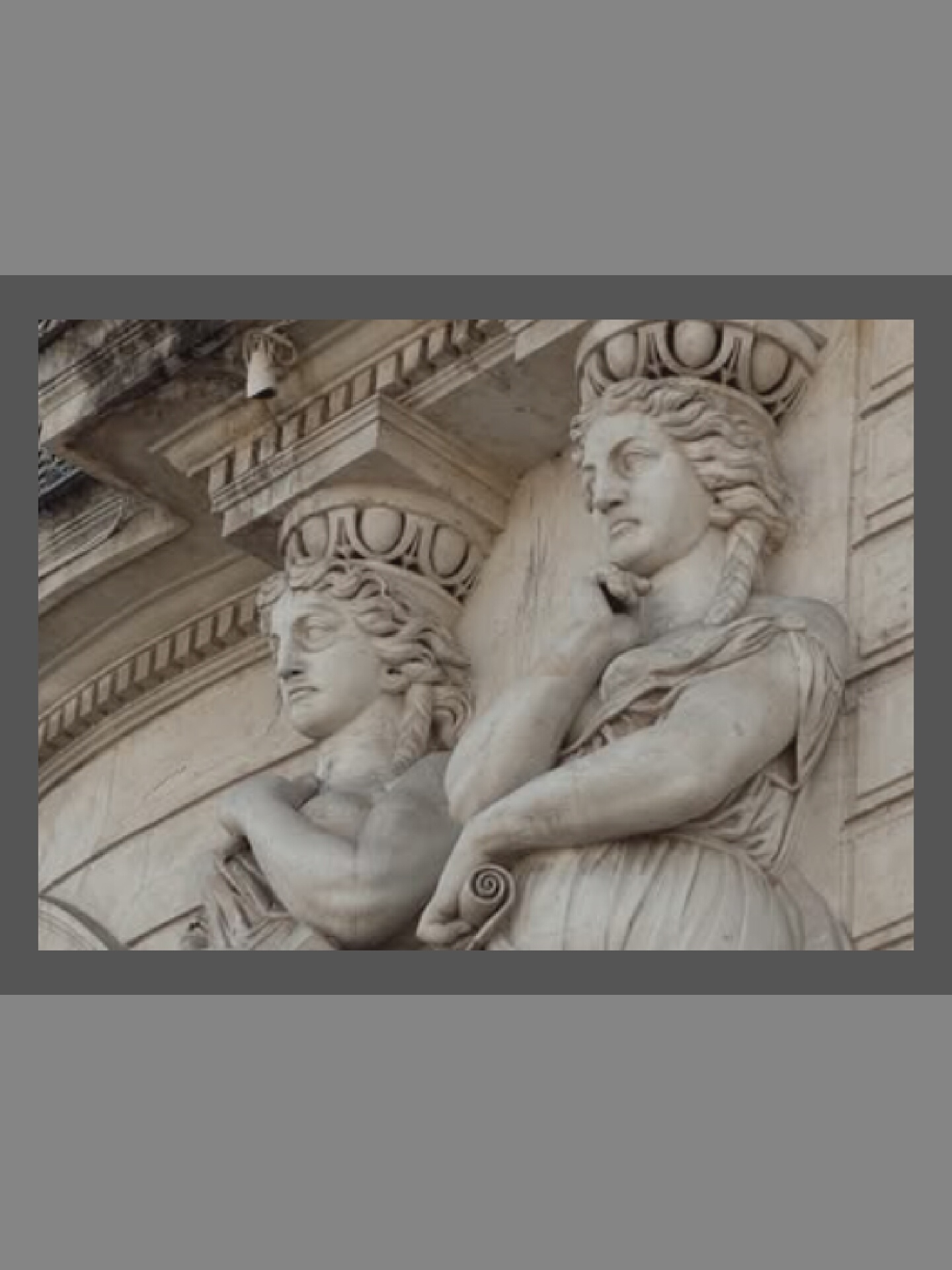 The image shows a close-up, eye-level, medium shot of two caryatids carved from white stone, supporting an architectural entablature. The caryatids, depicted as female figures in classical drapery, are positioned in profile, facing left. The caryatid on the right is more fully visible, with her head turned to the left, her gaze directed downwards. Her right arm is bent and raised, with her hand resting near her chin, while her left arm is bent across her chest, holding a scroll. The caryatid on the left is partially obscured by the figure on the right, but her head is also turned left, and her left arm is visible, bent at the elbow. The architectural elements above the caryatids feature decorative carvings, including what appears to be a volute on the left. The background is a light-colored, textured wall. The lighting is even, with no harsh shadows, suggesting an overcast day or an indoor setting. The image appears to be a detail from a building's facade or interior.