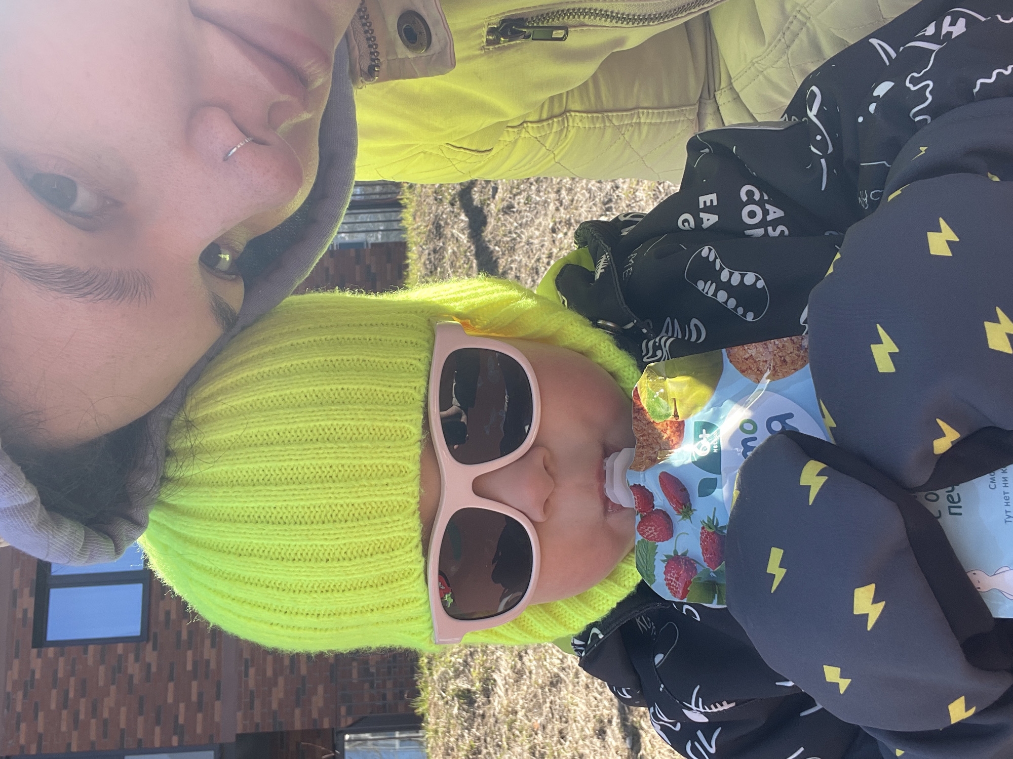 A vertically oriented, medium shot depicts two individuals. The upper left corner shows a woman from the shoulders up, wearing a light green zip-up jacket and a gray hooded sweatshirt. Her face is partially visible, with a nose ring.
Below and slightly to the right, a young child is featured, wearing a neon yellow beanie and oversized pink sunglasses. The child is drinking from a baby food pouch with strawberries and apples illustrated on the packaging. The child is wrapped in a black jacket with yellow lightning bolt patterns.
The background shows a building facade with brick-like patterns, small windows, and patches of dead grass. Based on the limited context, the specific location within the city of Izhevsk, Russia, cannot be definitively determined. The time of day is likely daytime due to the lighting conditions.