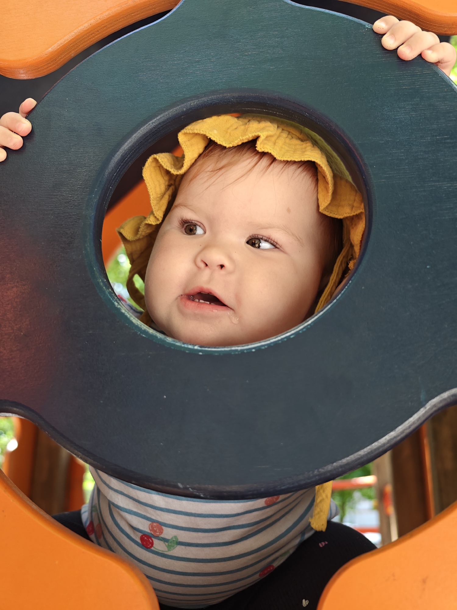 A close-up, eye-level shot shows a baby wearing a yellow bonnet and a bib with a blue and white striped pattern adorned with cherries. The baby is peeking through a circular opening in a blue wooden frame, which is part of an orange playground structure. The baby's hands are visible grasping the blue frame from the outside. The background is blurred, showing hints of greenery and what appears to be other playground elements, suggesting an outdoor setting. The image was captured on a sunny day, evident from the bright lighting. The specific location of this playground in Chaikovsky, Russia is unknown.