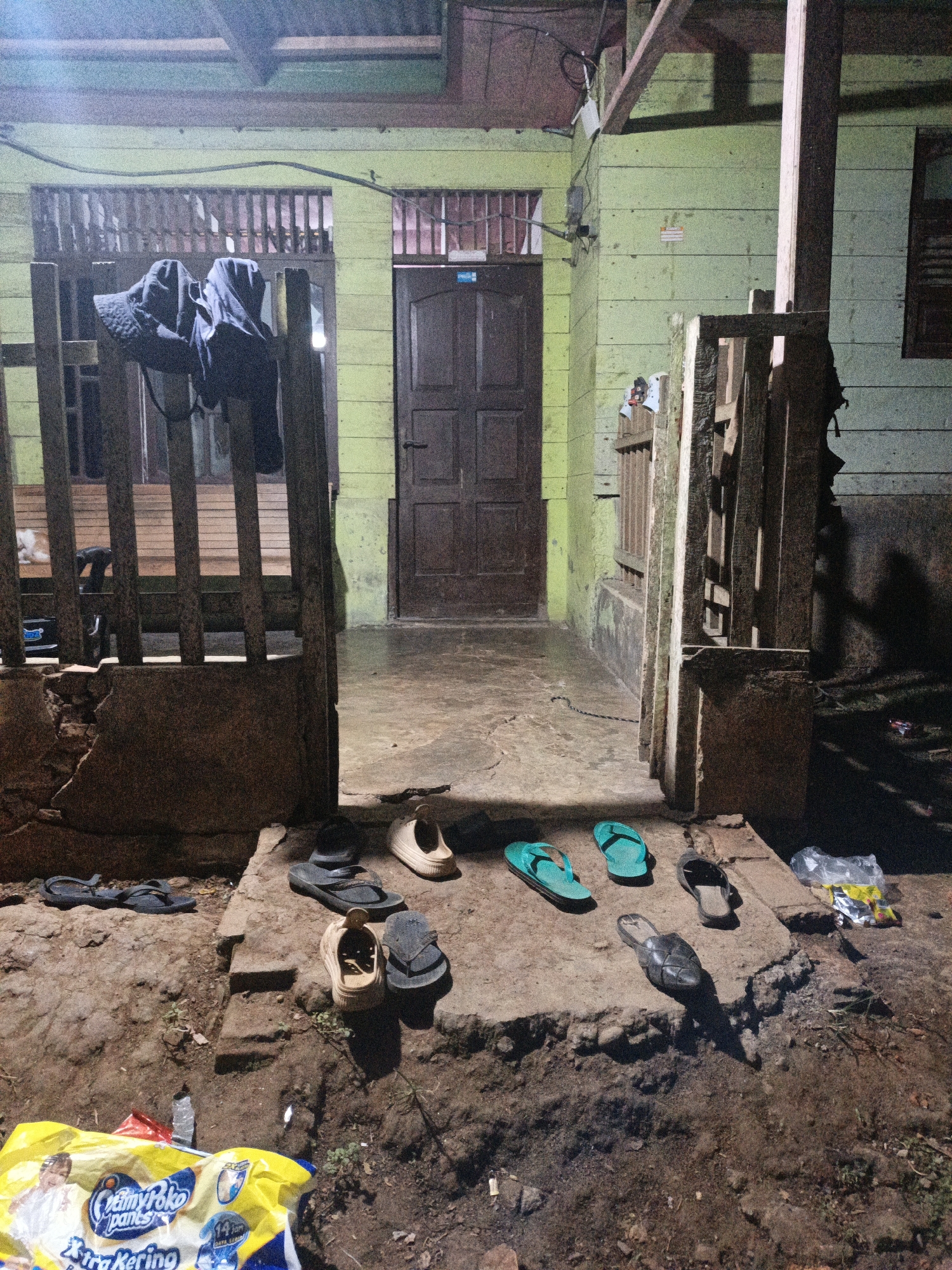 The image depicts the entrance to a simple, weathered wooden building in Meunasahuket, Indonesia. The scene is set at dusk or night, indicated by the artificial lighting casting shadows and the muted colors. A wooden porch with a railing extends to the left, with two dark-colored jackets or clothing items hung over it.
In front of the building's entrance, which features a dark wooden door, several pairs of footwear are scattered on a concrete pathway and the surrounding dirt. These include flip-flops in teal and dark colors, casual sandals, and a pair of light-colored slip-on shoes. The ground is uneven and appears to be a mix of dirt and worn concrete.
To the right of the entrance, another section of wooden fencing stands, and a dark shadow is cast on the light green wooden wall of the building. In the foreground, a bright yellow plastic bag is partially visible, with some text printed on it, including "CreamyPoko pants" and "Extra Kering". The overall impression is one of a humble, lived-in dwelling, likely in a rural or semi-rural area, with everyday items left outside. There are no people visible in the image.