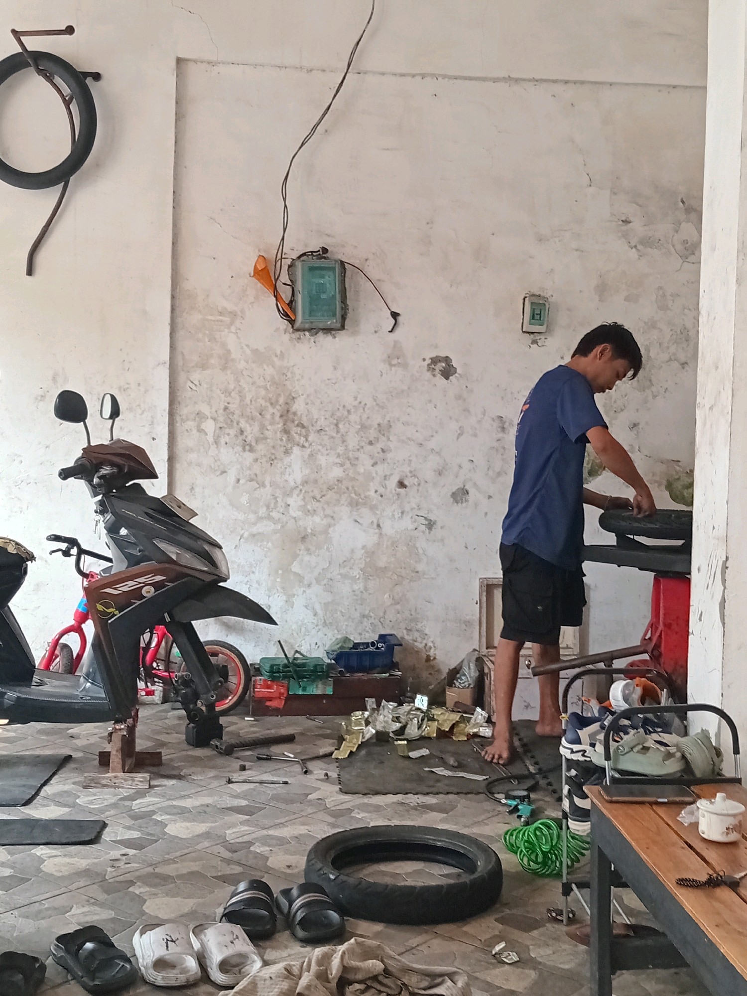 The image shows a man working on a motorcycle tire in a workshop, possibly in Tebalo, Indonesia. The man, wearing a blue t-shirt and black shorts, is seen standing and leaning over a tire mounted on a stand. The workshop appears to be an open-air or semi-enclosed space with a concrete floor and textured walls. Various motorcycle parts, tools, and a spare tire are scattered around the floor. A scooter is parked in the background, and a red children's bicycle is visible beside it. The scene suggests a typical day at a motorcycle repair shop.