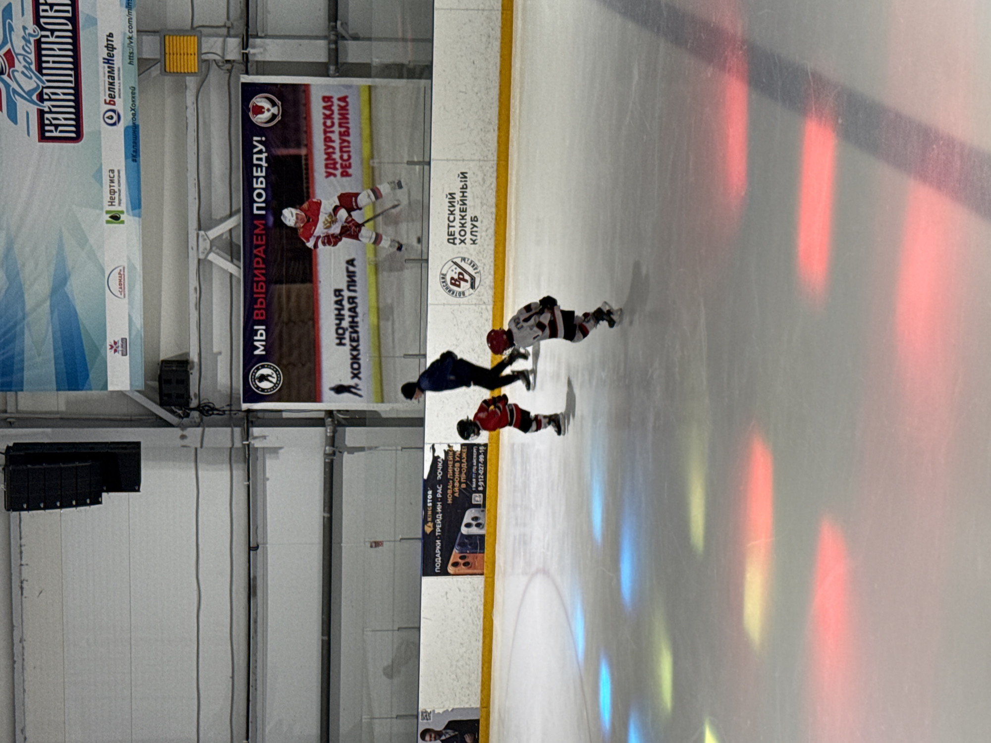 The image shows an indoor ice rink in Votkinsk, Russia, with a hockey game in progress. Three young children are visible on the ice, dressed in hockey gear. Two of the children are on skates, one in a red and white uniform, and the other in a white and black uniform. The third child, also in a white and black uniform, is facing away from the camera and skating towards the left. A partially visible adult figure in dark clothing is standing on the ice to the right of the children. The background features advertising banners, one of which displays "КУБОК КАЛАШНИКОВ" and "УДМУРТСКАЯ РЕСПУБЛИКА". The ice surface is illuminated by colorful light projections, primarily red, yellow, and blue. In the upper portion of the image, large black speakers are mounted on the ceiling.