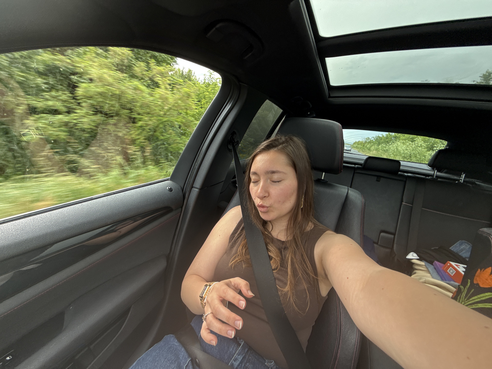 A young woman takes a selfie from the passenger seat of a moving car, likely in Bratovo, Bulgaria. She has brown hair, is wearing a brown sleeveless top and blue jeans, and is secured by a black seatbelt. Her eyes are closed, and her lips are slightly pursed in a pout. She wears a golden watch and rings on her left hand, which is positioned in front of her.
The car's interior features black leather seats with red stitching and a black ceiling with a large, open panoramic sunroof, revealing an overcast sky. Through the blurred side windows, a lush green, tree-filled landscape indicates the car is traveling at speed. A faint BMW logo is visible on the passenger window. Various personal items, including clothing, a bag with an orange floral pattern, and an indistinct red and white box, are piled on the back seat. The scene suggests a daytime journey under cloudy weather conditions.