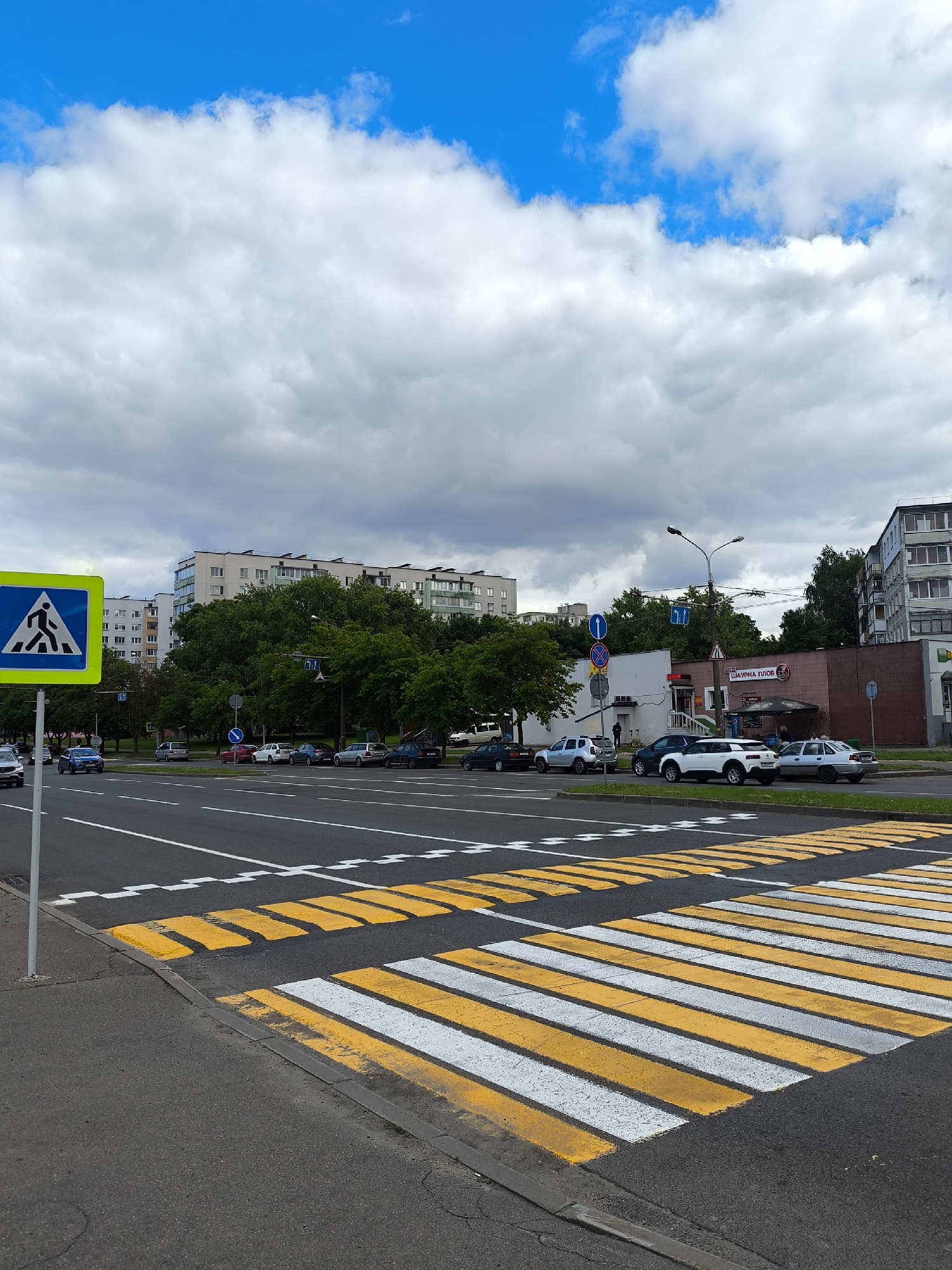 A sunny day with some clouds over a street in Minsk, Belarus. In the foreground, a pedestrian crossing with yellow and white stripes is visible. A blue and yellow pedestrian crossing sign is on the left side of the image. The street is lined with cars, trees, and buildings in the background. A few people are visible walking on the sidewalk near a building with a sign that reads "Шаурма Плов".