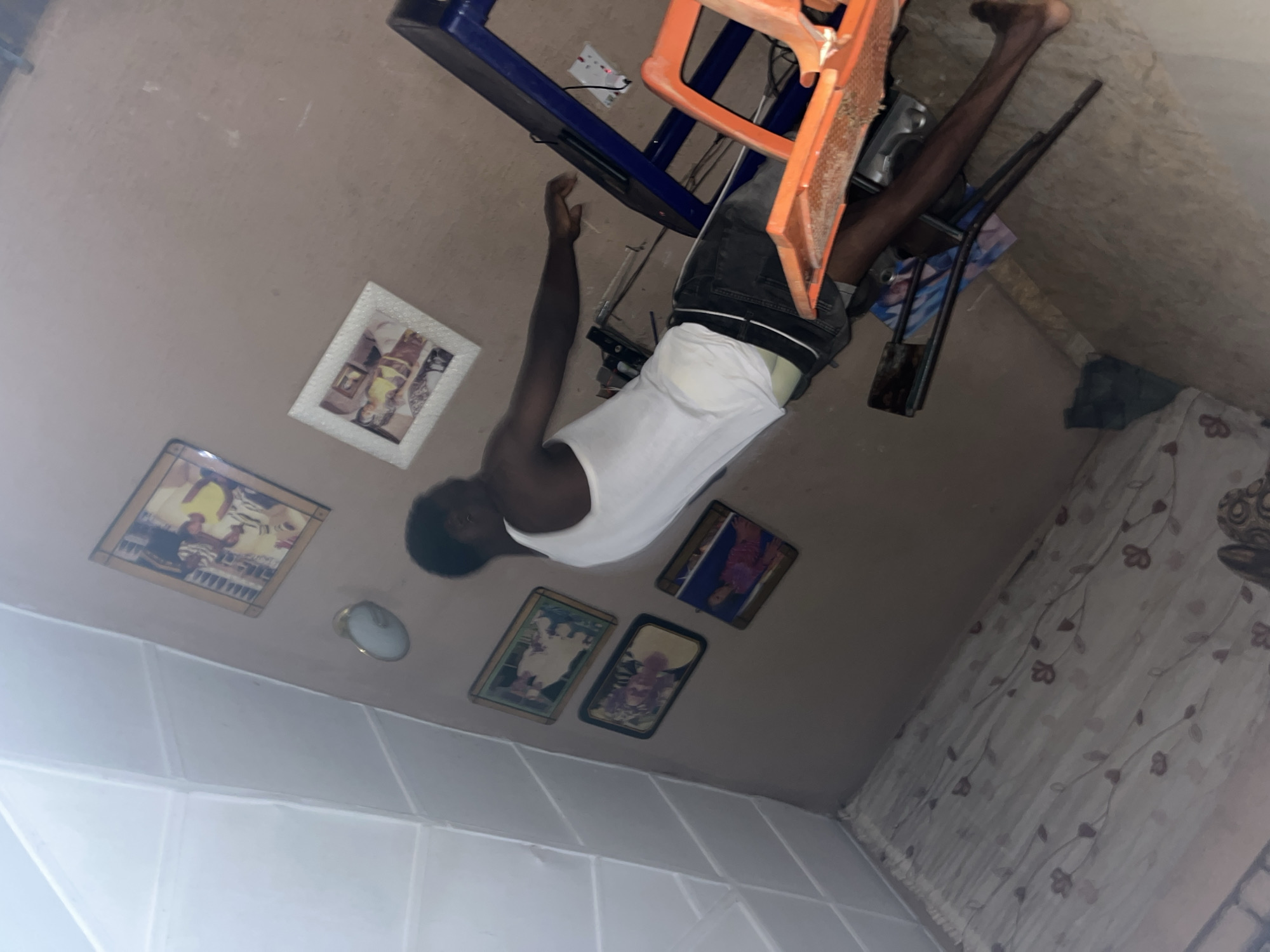 The image, taken in an indoor environment of unknown location, depicts a young Black male standing near a treadmill. He is wearing a white tank top and dark denim shorts, with a white boxer brief visible at the waistline. His left arm is extended, and his right arm is bent at the elbow. The room's walls are a neutral brown color, and the ceiling is white with geometric patterns. Several framed pictures hang on the wall. To the right of the male is a blue treadmill with an orange plastic seat attached. Electrical outlets are visible on the treadmill's frame. The floor area shows a patterned curtain in the foreground, and part of a wooden surface in the background. The lighting is dim, with a visible overhead light fixture on the wall.