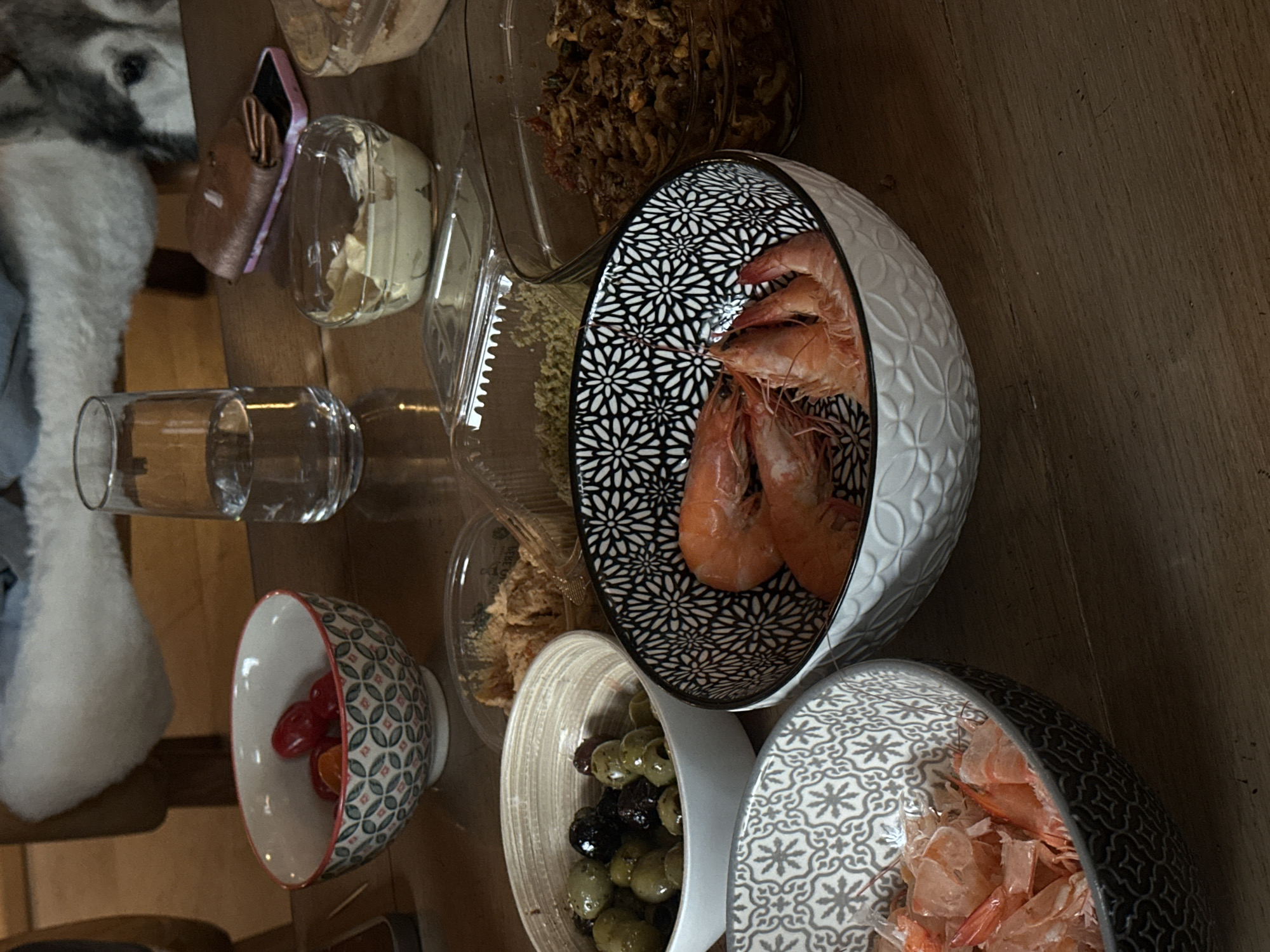 The image shows a wooden table laden with various food items and containers, likely in a domestic setting. There are bowls containing shrimp, olives, and cherry tomatoes. Additionally, there are clear containers holding spreads, possibly pate or dip, and a jar of fried onions. A glass of water and a purse are also present. A portion of a white dog is visible in the left side of the frame. Location in the city of La Couarde-sur-Mer, France is not evident from the image.