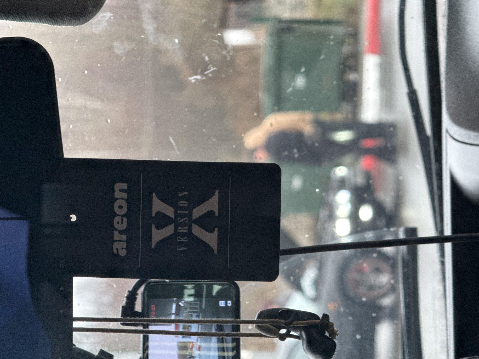 The image provides a view from inside a vehicle, looking out through a windshield that appears wet and somewhat dirty. The original image is oriented vertically, showing elements both inside and outside the vehicle.
In the foreground, hanging inside the car, is a black air freshener with the brand "areon" and "VERSION X" clearly visible in white text. Below it, a smartphone is mounted, likely functioning as a dashcam or recording device, with its screen displaying the time "02:23". A small, dark, perhaps decorative, object hangs by a cord to the right of the phone. Part of the vehicle's interior, possibly a sun visor, is visible at the top left.
The outdoor scene, viewed through the rain-streaked glass, appears blurry and dimly lit, suggesting overcast conditions or low light, consistent with the early morning time shown on the phone (02:23). In the mid-ground, a red and white striped barrier or pole is visible on the right, indicating a possible checkpoint or restricted area. Green dumpsters or containers are visible further in the background.
A dark-colored car with distinctive circular headlights is seen from the rear in the center-right of the frame. Behind this vehicle, a person wearing a light-colored top is bent over, their activity unclear due to blur and obstruction. Another person, dressed in darker clothing, is partially visible nearby. The scene suggests the vehicle from which the photo is taken is stopped or waiting.
The visible text includes "areon", "VERSION X", and the digital time "02:23". The location context for this scene is Klenovskoe, Russia.