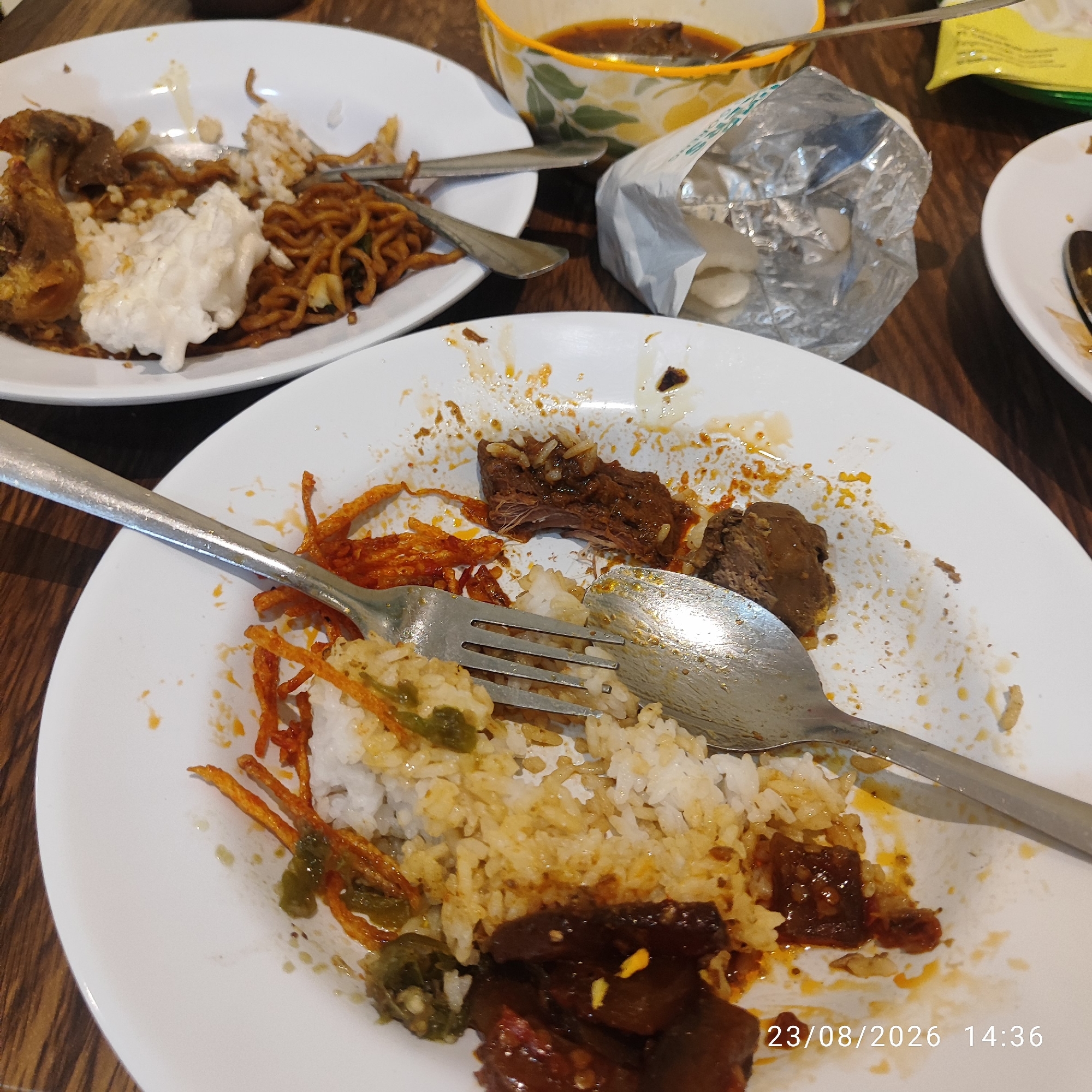 The image depicts a messy, half-eaten meal on a white plate, likely at a restaurant or dining establishment. The meal appears to consist of rice, noodles, and pieces of meat, with remnants of sauce and garnish scattered across the plate and the surrounding wooden table. A fork and spoon are present, indicating recent consumption. In the background, other plates with food remnants, a bowl of sauce, and a crumpled foil wrapper are visible. The overall scene suggests a casual dining experience where the meal is nearing completion. The date and time stamp "23/08/2026 14:36" is superimposed on the image, but the location in Tanjung Pendam, Indonesia, is not explicitly visible or inferable from the image content.