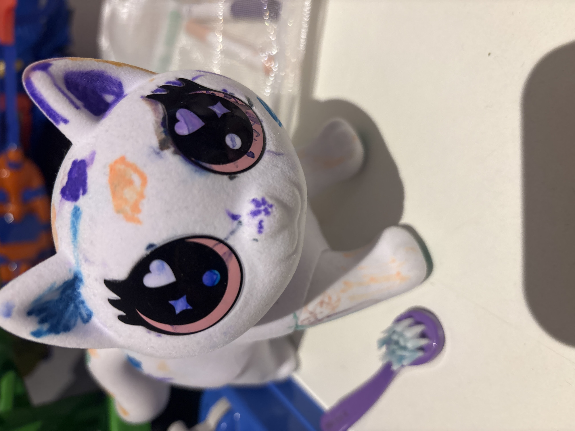 The image displays a white, cat-shaped plush toy or figurine positioned centrally on a light-colored surface. The toy is decorated with various smudged or dabbed colors, including purple, blue, and orange, across its body and ears. Its large, cartoonish eyes are black with distinct pink and purple outlines, featuring white heart-shaped pupils and smaller blue or purple star-like reflections. A small purple star-shaped smudge is visible below the toy's left eye. To the left of the cat, a small, purple child's toothbrush with light blue bristles rests on the same surface. The background is blurred, revealing indistinct colorful objects, primarily in shades of blue, orange, and green, suggesting an indoor setting with other toys or items. A blue tray or container is partially visible on the far left. No individuals are present. The precise location within the city of Dubai, United Arab Emirates, cannot be determined from the visual information provided.