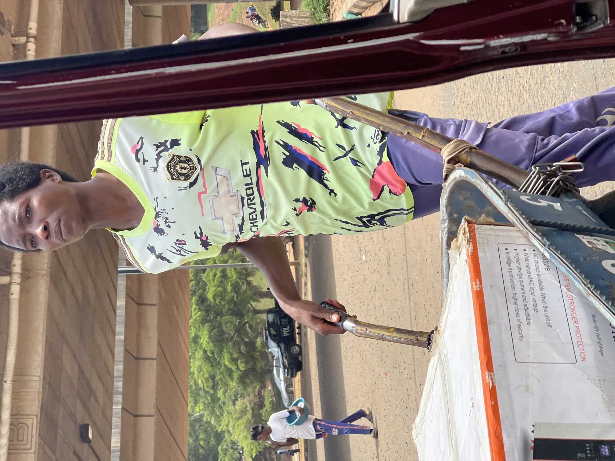 The image shows a man in a Manchester United Chevrolet soccer jersey and purple pants operating a tricycle in what appears to be Abuja, Nigeria. He is holding the handlebars and looking toward the camera. The tricycle is carrying a box with the text "SYSTEM STRONGER PROTECTION," likely a commercial product. In the background, a police car is visible on the road, and another man in a white shirt and purple pants is walking along the street. The scene is set under a bridge, with the support columns visible on the left and greenery on the right.