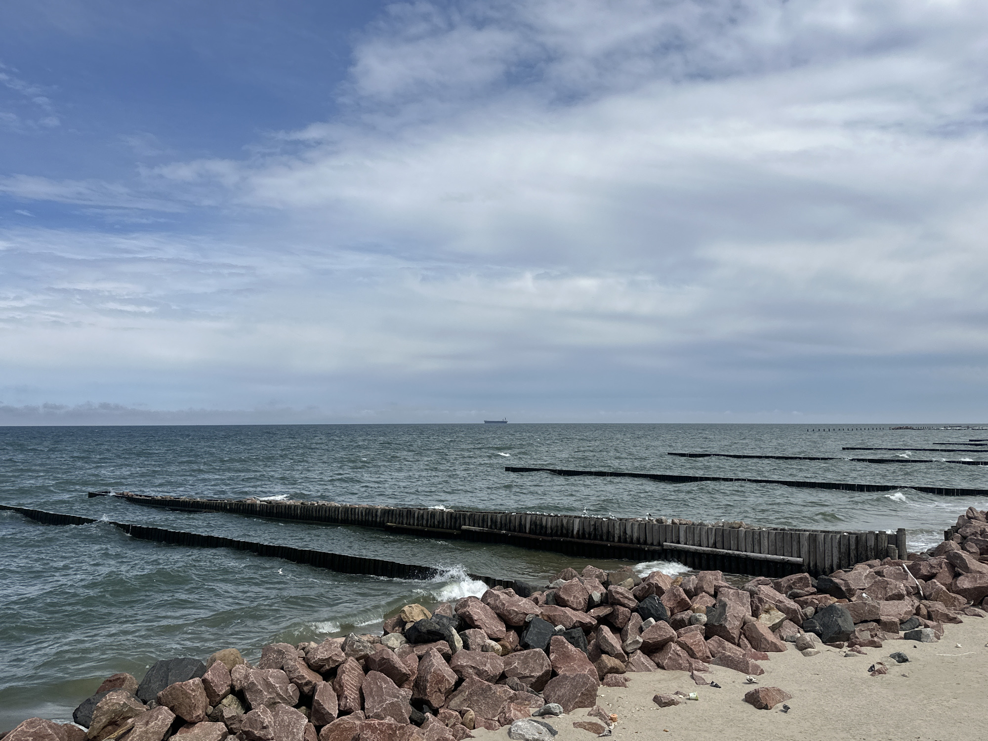 The image shows a wide view of the Baltic Sea near Svetlogorsk, Russia. In the foreground, a sandy beach is covered with large, rough-cut pink and gray rocks. A line of wooden breakwaters stretches out into the dark, choppy water, with waves breaking against them. Several more lines of breakwaters are visible further out into the sea, creating parallel patterns. In the distance, a large cargo ship is visible on the horizon under a sky filled with a mix of white and gray clouds. The scene is a natural coastal landscape with man-made structures for erosion control.