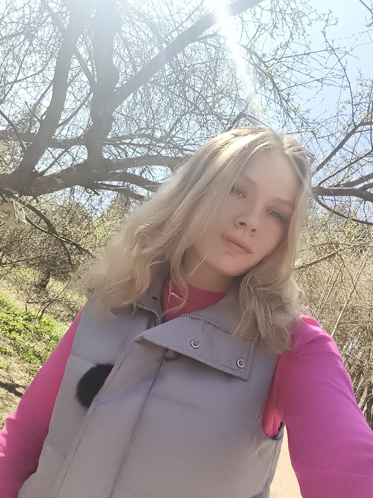 A young woman with blonde hair and fair skin is posing for a selfie outdoors. She is wearing a bright pink long-sleeved shirt and a light gray puffer vest. A small, dark, round object is attached to her vest, possibly a microphone or decorative item. The background is a tree with bare branches against a blue sky with bright sunlight causing lens flare. The location appears to be a park or wooded area in the daytime. There is green vegetation visible in the lower left corner. The image angle is slightly upward, suggesting the woman is holding the camera above her head. The location is Izhevsk, Russia.