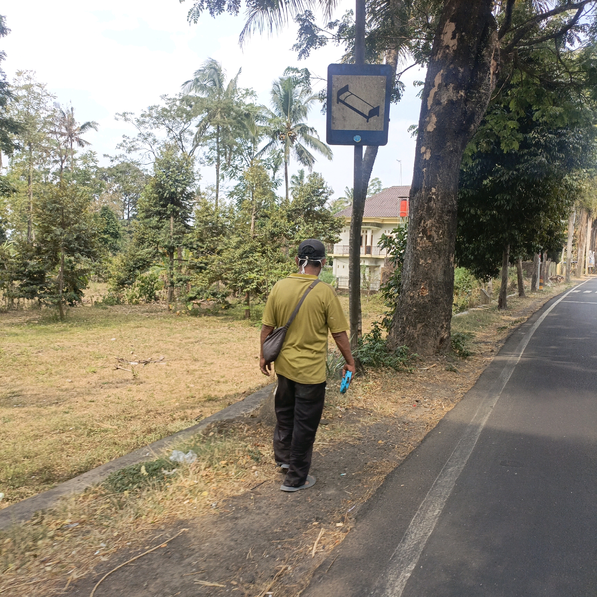 A person wearing a yellow t-shirt, black pants, a black cap, and a face mask walks along a paved road in Salatiga, Indonesia. The person carries a dark brown shoulder bag and a blue object in their right hand. To the left of the road is a grassy area with sparse trees and vegetation. A large tree stands near the road, and a sign with a bed symbol is mounted on a pole beside it. In the background, a white house with a brown roof is visible, surrounded by trees. The sky is cloudy.