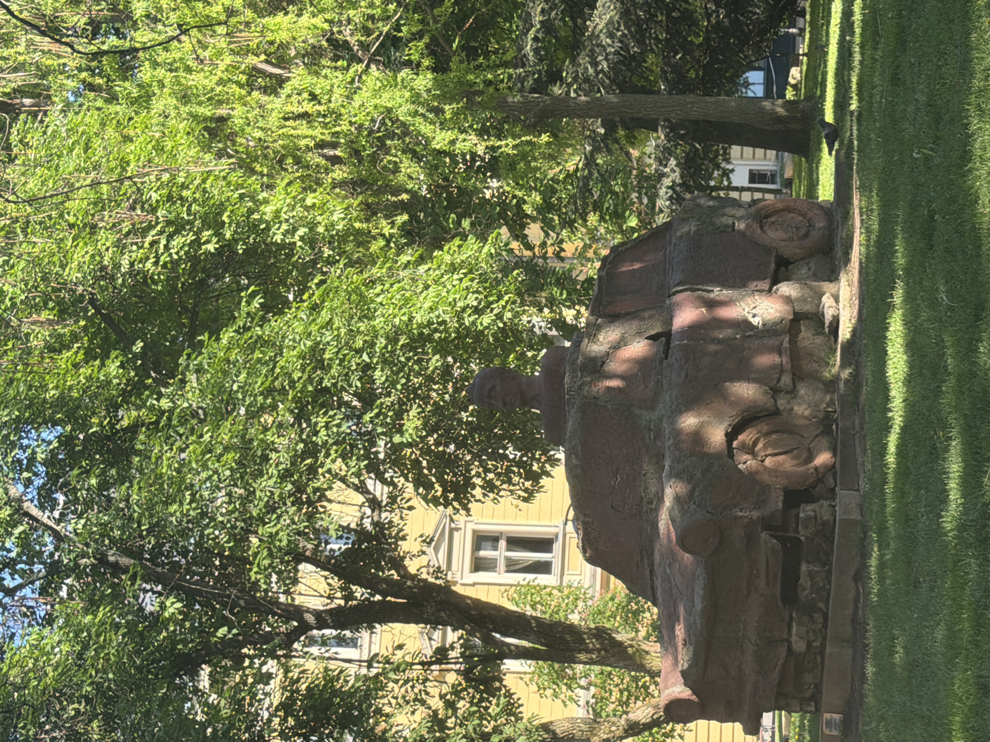 The image shows the Monument to the Victims of Communism in Sofia, Bulgaria. The statue depicts a crushed car with figures emerging from the rubble. It is located in a park setting with trees and a building visible in the background. A pigeon is seen on the grass in the upper right. The image is taken from a ground-level perspective.
