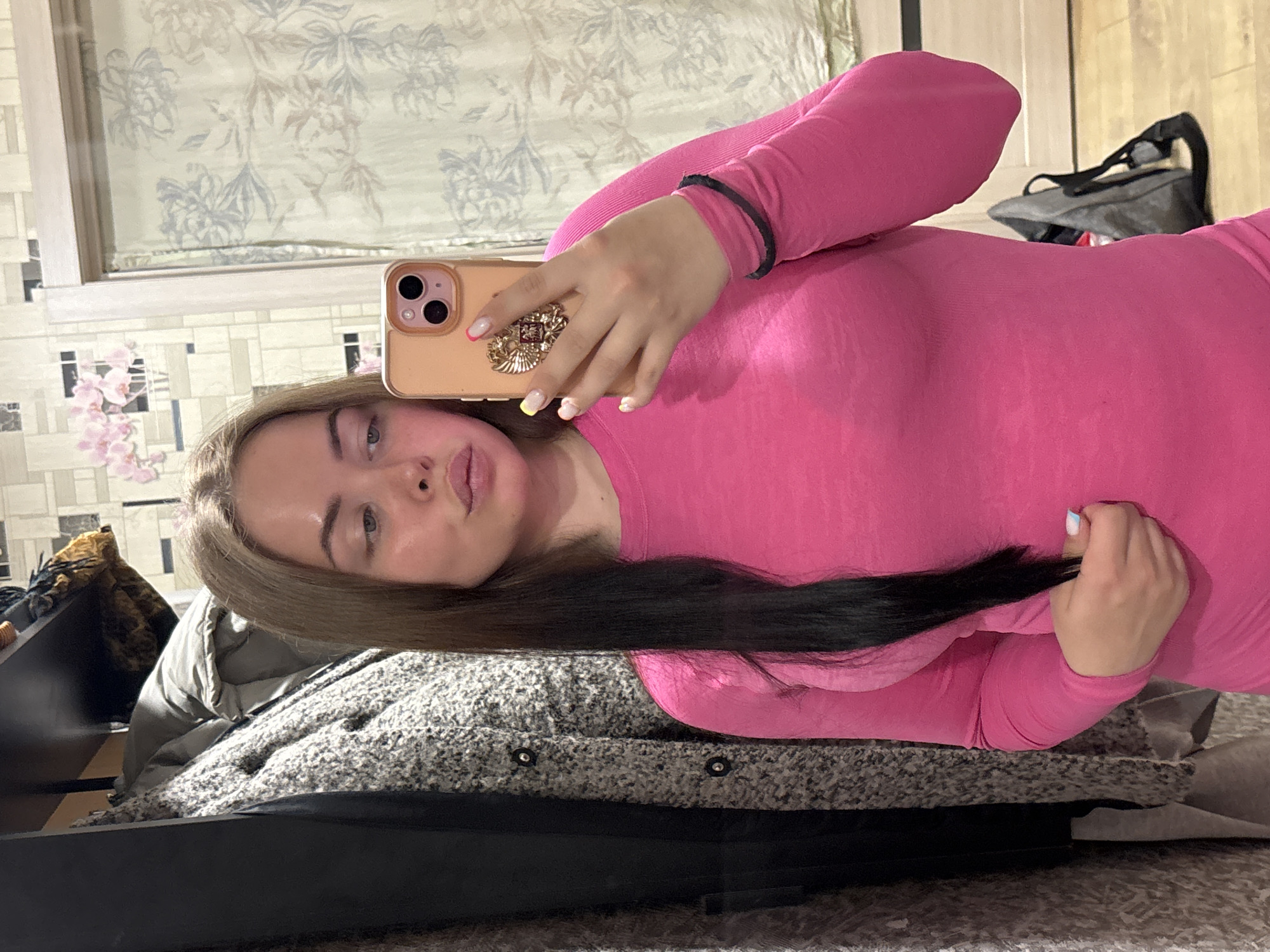 A young woman is taking a selfie in a room. She has long, two-toned hair that is blonde at the roots and dark brown at the ends. She is wearing a bright pink, long-sleeved top and holding a peach-colored smartphone with a decorative element on the back. She is looking directly at the camera.
The room appears to be a bedroom, based on the presence of a bed with patterned bedding and a gray, textured headboard. There's also a curtained window and a tiled wall with floral decorations. A gray bag is visible in the background.
It is difficult to ascertain the location in Izhevsk, Russia, from this image alone. Additional information about identifiable landmarks or distinct architectural elements would be required.
