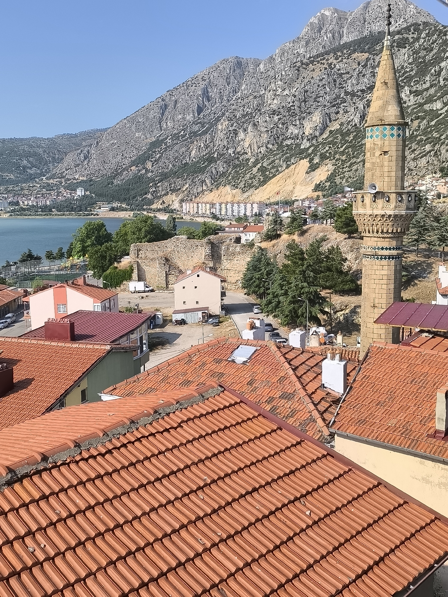 The image captures a panoramic view of the city of Eğirdir, Turkey, with a prominent minaret of a mosque in the foreground, set against a backdrop of a rugged mountain range and a calm body of water, likely Eğirdir Lake. The foreground is dominated by a dense cluster of terracotta-tiled roofs, showcasing the architectural style of the region. Several residential buildings with varying colors and designs are visible amidst the rooftops. Further into the scene, the clear blue water of the lake stretches across the horizon, with a few boats dotting its surface. On the opposite bank, a town with a mix of modern and older buildings is nestled at the foot of the imposing, rocky mountains. The slopes of these mountains are sparsely vegetated, with some areas showing evidence of erosion. The sky is a clear, bright blue, suggesting a sunny day. The overall impression is one of a picturesque and tranquil lakeside city, characterized by its natural beauty and architectural heritage.