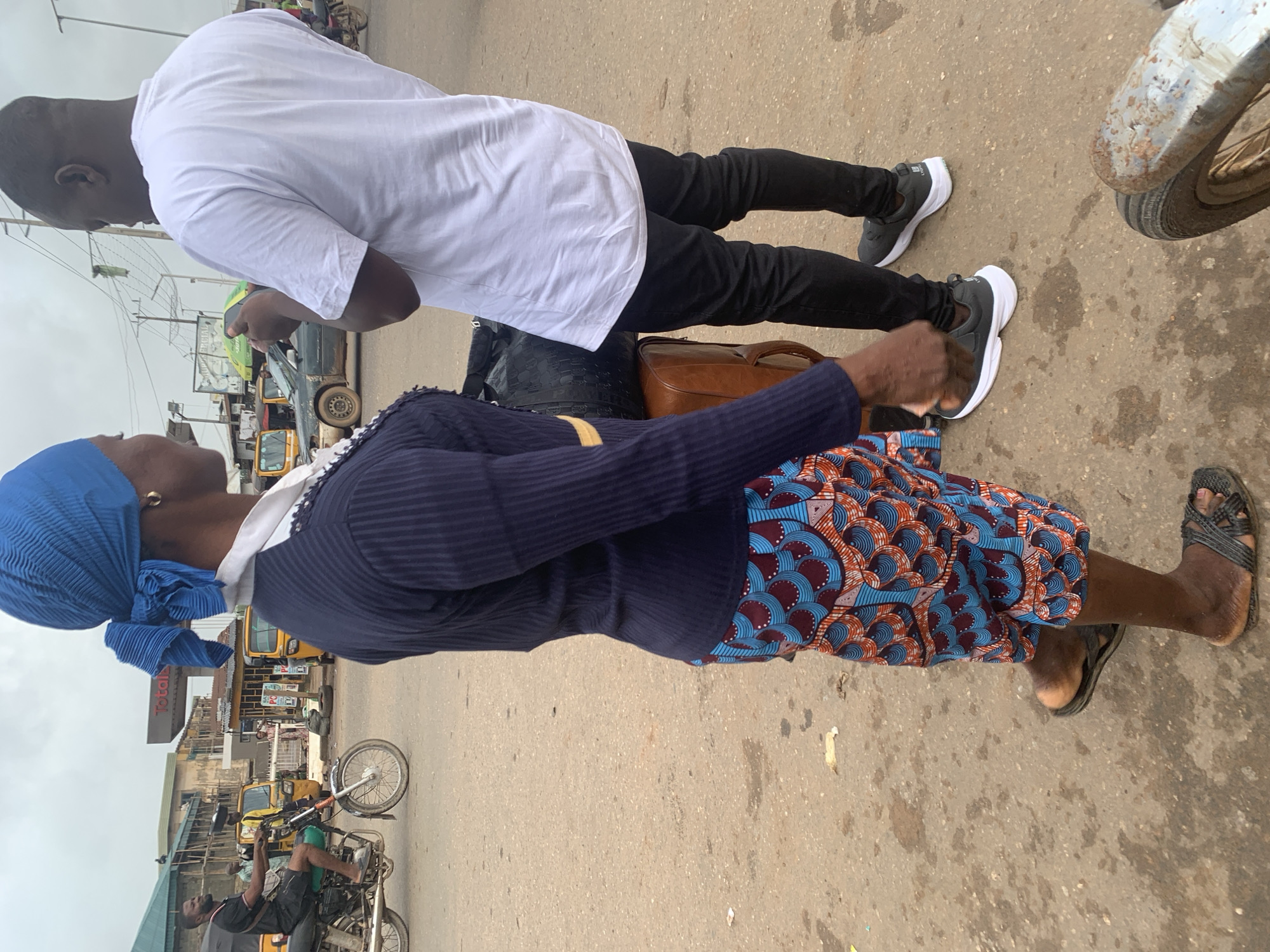 The image shows a street scene in Shagamu, Nigeria, with two people in the foreground. The person on the left is a woman wearing a blue headscarf, a dark blue ribbed sweater with a white collar, and a colorful patterned skirt. She is also wearing dark sandals. She is standing next to a brown suitcase and a black duffel bag. The person on the right is a man wearing a white t-shirt and black pants. He is looking down at his phone.
In the background, there are several vehicles, including a motorcycle with a rider, several yellow auto-rickshaws (keke napep), and a dark-colored car. There are buildings and utility poles visible, suggesting an urban environment. The ground is paved with asphalt and appears to be a street or market area. The sky is overcast.