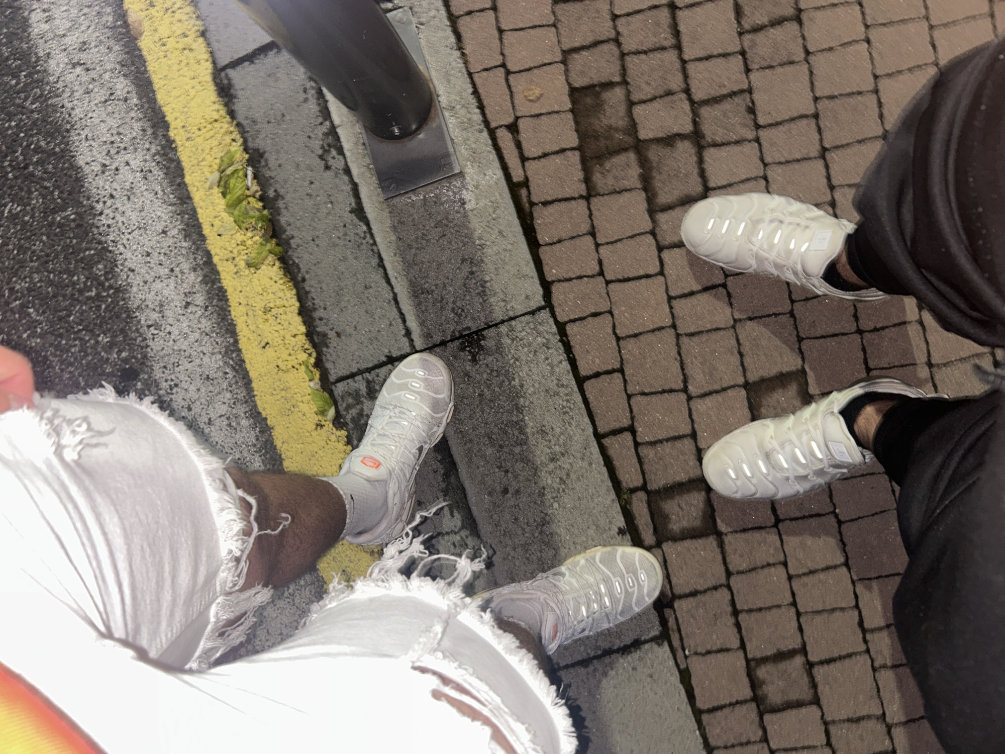 An overhead shot captures the legs and feet of two individuals standing on an urban or semi-urban surface at night. One person, positioned on the left, wears light-colored (likely white) ripped jeans, white socks, and white sneakers. The sneakers are identifiable as Nike Air VaporMax Plus, featuring an orange "TN" logo on the tongue and "VAPORMAX PLUS" text on a side tag. The other person, on the right, wears dark pants and similar white Nike Air VaporMax Plus sneakers with a visible side tag.
The ground comprises multiple textures: dark asphalt with a prominent yellow painted line running vertically, a concrete curb separating the asphalt from brick pavers, and brick pavers on the right side of the image. Small patches of green debris, possibly leafy greens, are visible on and near the yellow line. A black pole, likely a street light or signpost, is anchored to the concrete curb. Several wet spots or small puddles are noticeable on the brick pavers, suggesting recent rain. The scene is illuminated by artificial light, reflecting off the sneakers and wet surfaces, indicating it is nighttime or very low light. The image captures a casual outdoor moment in Dzhebel, Bulgaria.