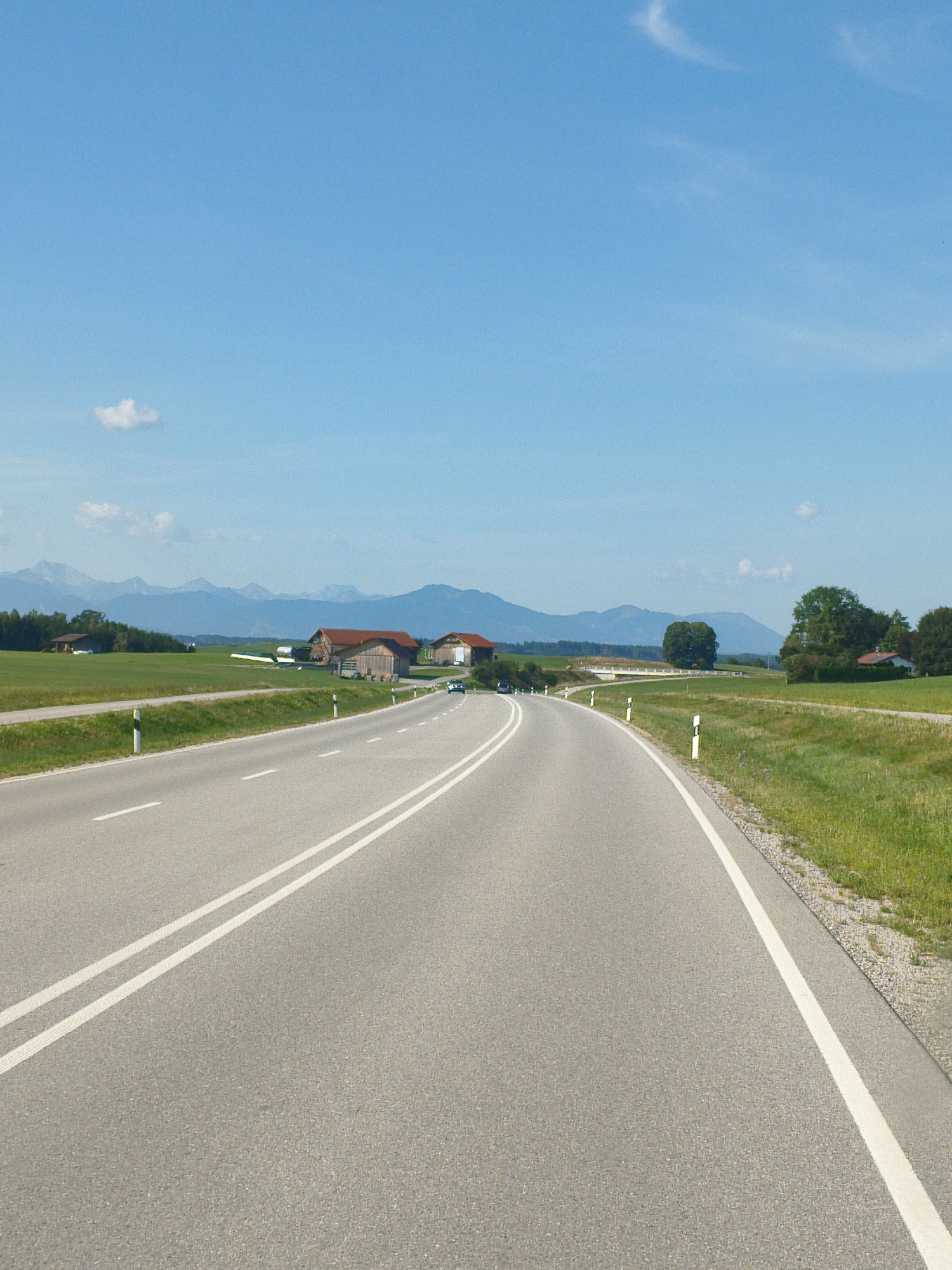 The image shows a rural road in Marktoberdorf, Germany. A two-lane asphalt road curves to the right. White lines mark the lanes and the edge of the road. On the left side of the road, there are guard posts with reflective markers. The landscape features rolling green hills, a few farm buildings with red roofs, trees, and distant mountains under a clear blue sky with a few scattered clouds. A dark-colored car is visible on the road in the middle distance, and another car is parked on the left side of the road near the guard posts. The environment appears to be a sunny day in the countryside.